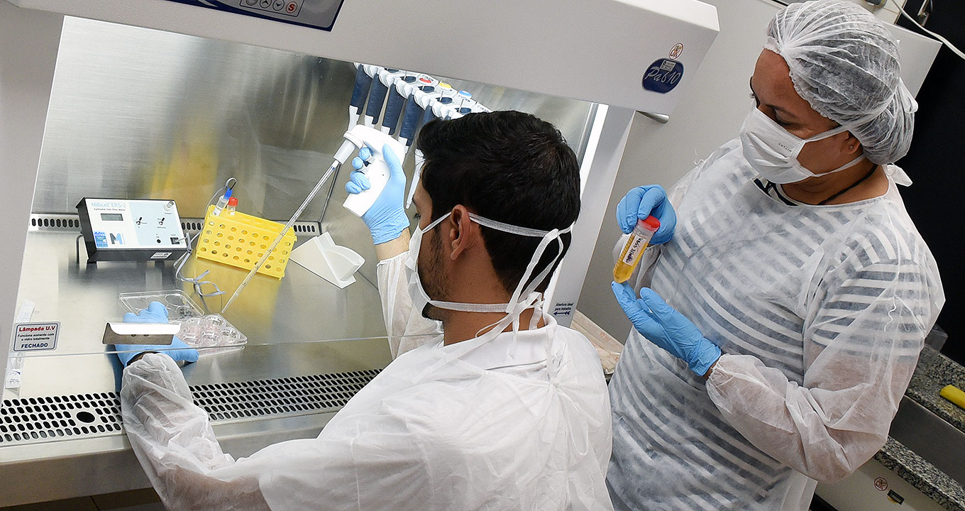 Em laboratório, imagem em perspectiva, de costas, e plano médio, homem sentado manipula substância com um tipo de dosador, usando uma bancada de inox que encontra-se à frente, com cerca de um metro de largura por sessenta centímetros de profundidade e que fornece luz da parte superior. Ele opera o dosador com a mão direita apontando a extremidade do equipamento para um tipo de coletor semelhante a uma forma de gelo que ele segura com a mão esquerda. À direita na imagem, mulher em pé observa-o. Imagem 1 de 1