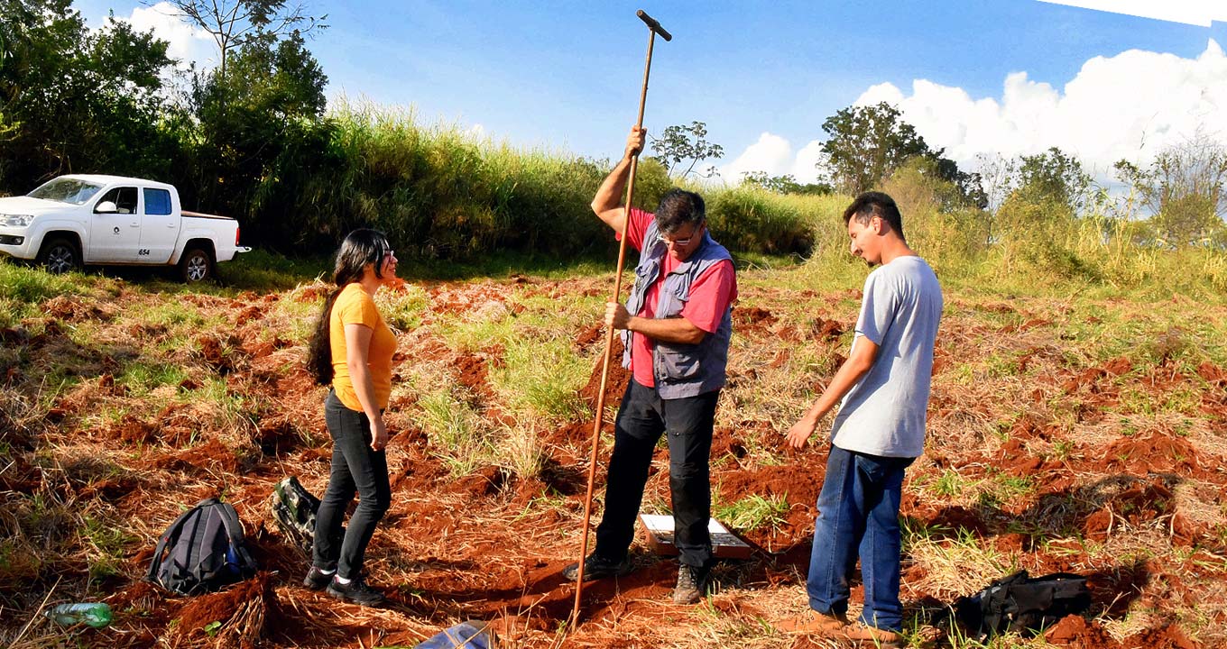 O professor Francisco Ladeira e alunos em trabalho de campo | Foto: Antoninho Perri Audiodescrição: Em campo aberto, área com vegetação rasteira capinada onde aparecem terra revolvida e mato seco, três pessoas em pé, sendo um homem ao centro, que perfura o solo com uma extensa vara de ferro, sendo observado por duas outras pessoas posicionadas junto a ele, à esquerda e à direita. Ao fundo há uma extensa área com vegetação alta e um veículo pick-up estacionado reste à esta vegetação. O céu está azul e a área ensolarada. Imagem 1 de 1.