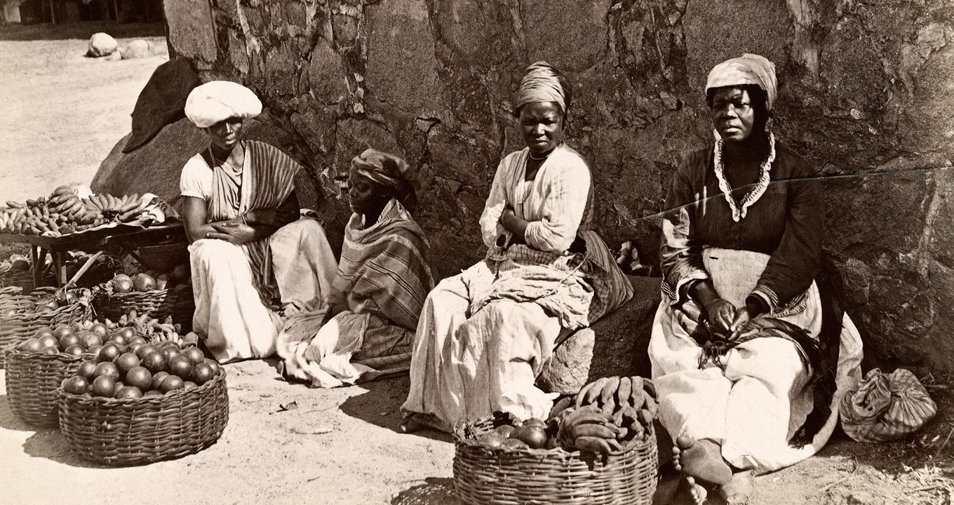 Audiodescrição: Em área externa, imagem em perspectiva e de corpo inteiro, em foto preto e branco de 1875, quatro mulheres negras na rua, sentadas no chão e sobre pedras, uma ao lado da outras. Elas vendem frutas como banana e abacates, que encontram à frente delas, dentro de seis cestos de vime, com cerca de oitenta centímetros de diâmetro. Às costas delas, há um paredão de pedras, que projeta sombra em algumas delas. Elas vestem saias e usam turbantes. Imagem 1 de 1.