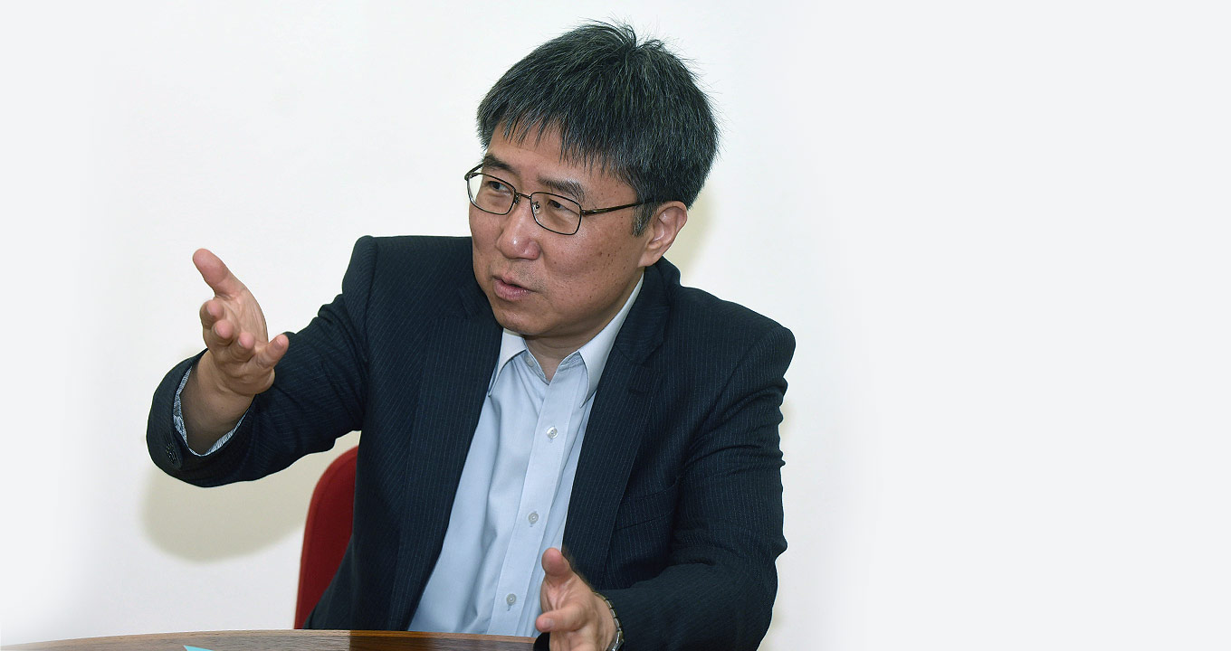 O economista Ha-Joon Chang: conferências lotaram o auditório do Instituto de Economia da Unicamp | Foto: Antonio Scarpinetti Audiodescrição: Em sala, imagem frontal e plano médio, homem sentado em cadeira fala gesticulando com o braço direito, mantendo-o erguido e levemente projetado para frente, e a mão aberta, com a palma voltada para o lado direito da imagem. O braço esquerdo permanece apoiado sobre uma mesa de madeira redonda que está à frente dele. Ele usa óculos e veste blazer preto sobre camisa social. Imagem 1 de 1.