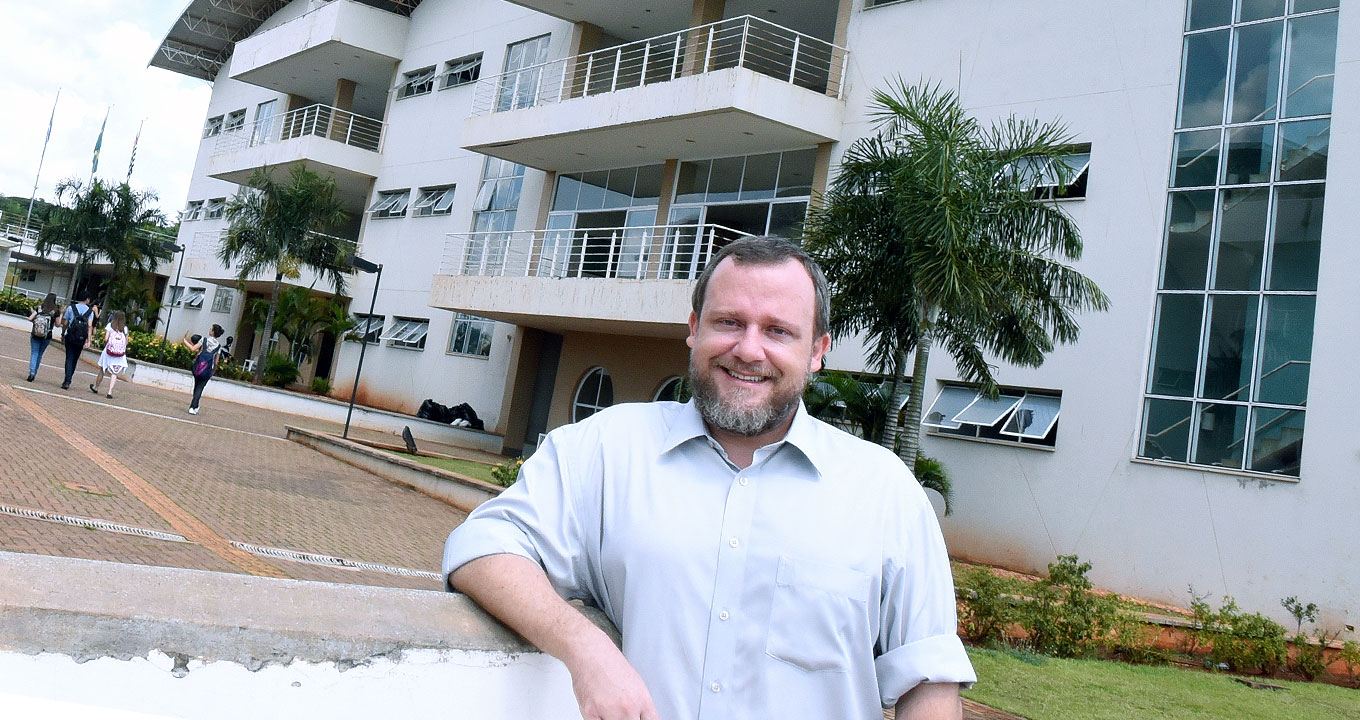 Renato Georgette Frigo, autor da pesquisa | Foto: Antoninho Perri Audiodescrição: Em área externa, imagem frontal e de busto, homem em pé, ao centro, sorri, sendo que mantém o braço esquerdo abaixado e o direito apoiado com o antebraço em uma mureta. Ele usa camisa de botões branca e de mangas compridas, que estão arregaçadas. Ao fundo, em perspectiva, à direita, uma edificação de três andares com sacadas e várias janelas e portas retangulares de vidro, com paredes pintadas de branco. Há palmeiras em frente ao prédio e amplo calçamento com piso intertravado. Imagem 1 de 1