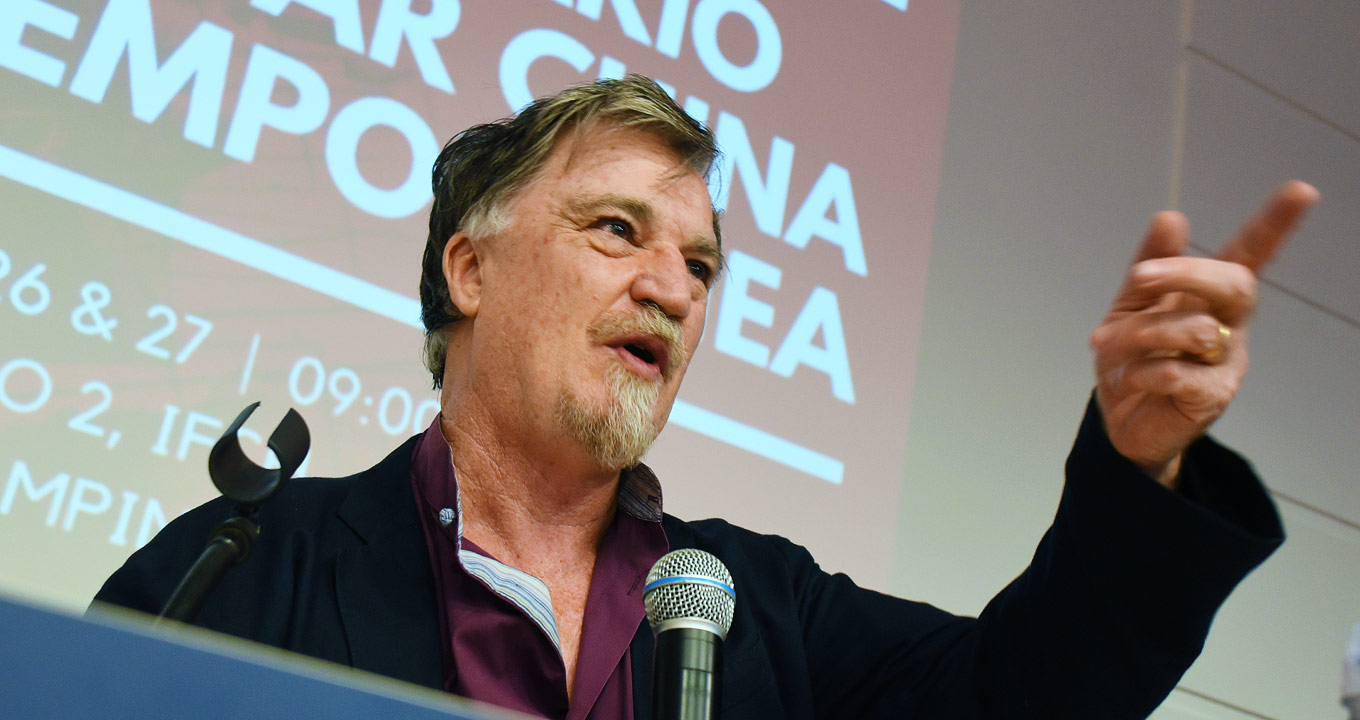 O professor e sociólogo Thomas Patrick Dwyer: pioneirismo nos estudos sobre a China | Foto: Antoninho Perri Audiodescrição: Em auditório, imagem em perspectiva e de busto, homem fala a microfone, com olhar voltado para a direita na imagem, sendo que mantém o braço esquerdo esticado para frente, apontando com o dedo indicador. Às costas dele, há uma grande tela de projeção de data show com frase escrita em letras brancas em fundo vermelho. Ele veste blazer azul marinho sobre camisa lilás. Imagem 1 de 1.