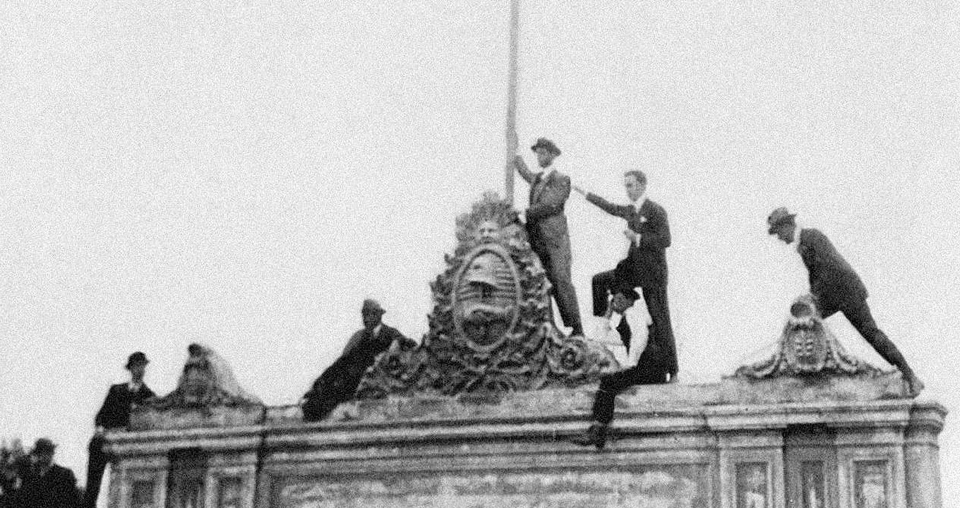 Audiodescrição: Em área externa, imagem frontal e de corpo inteiro, em foto em preto e branco antiga, datada de 1918, cerca de sete homem no topo de uma edificação, sendo que alguns encontram-se sentados e outros em pé. Todos vestem roupas sociais, como terno, e chapéu. Imagem 1 de 1.