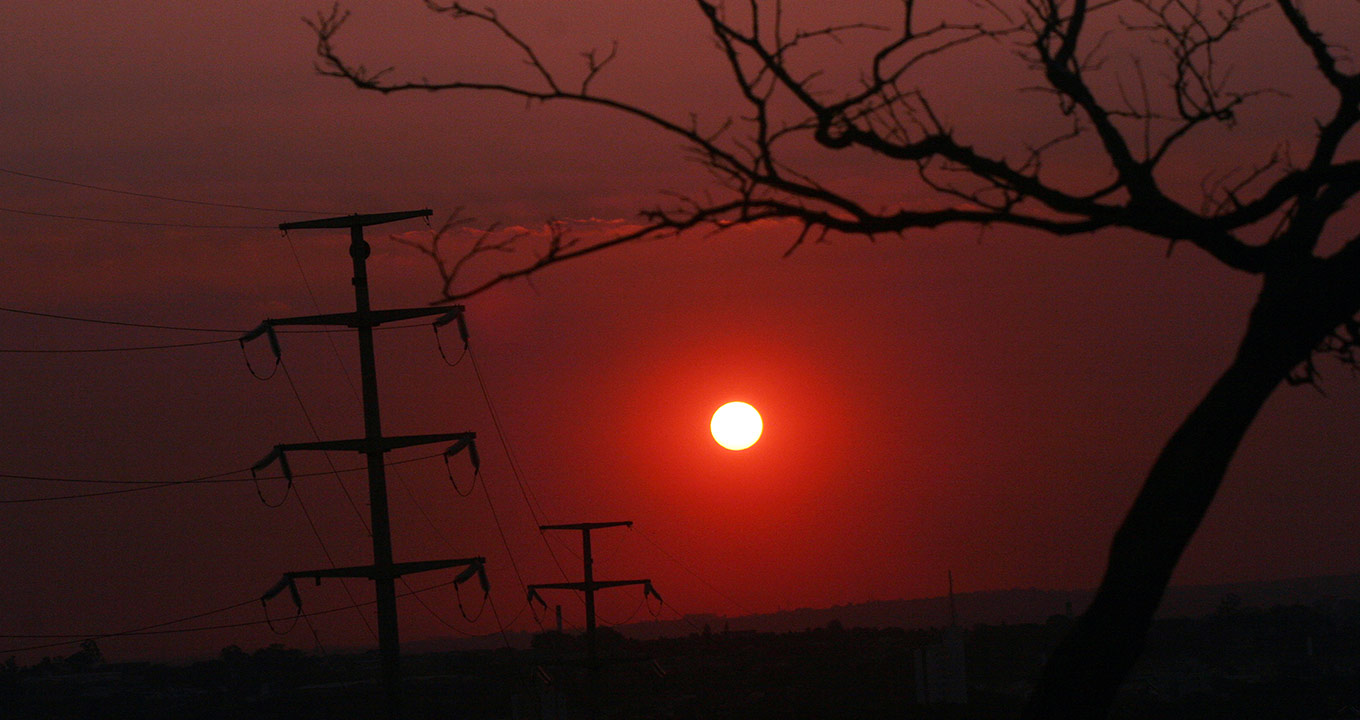 Audiodescrição: Em área externa, imagem de pôr-do-sol, bastante escura e alaranjada, sendo que à frente, a direita na imagem, há galhos e tronco de uma árvore, e à esquerda, altas torres com fios de transmissão de energia. Imagem 1 de 1.