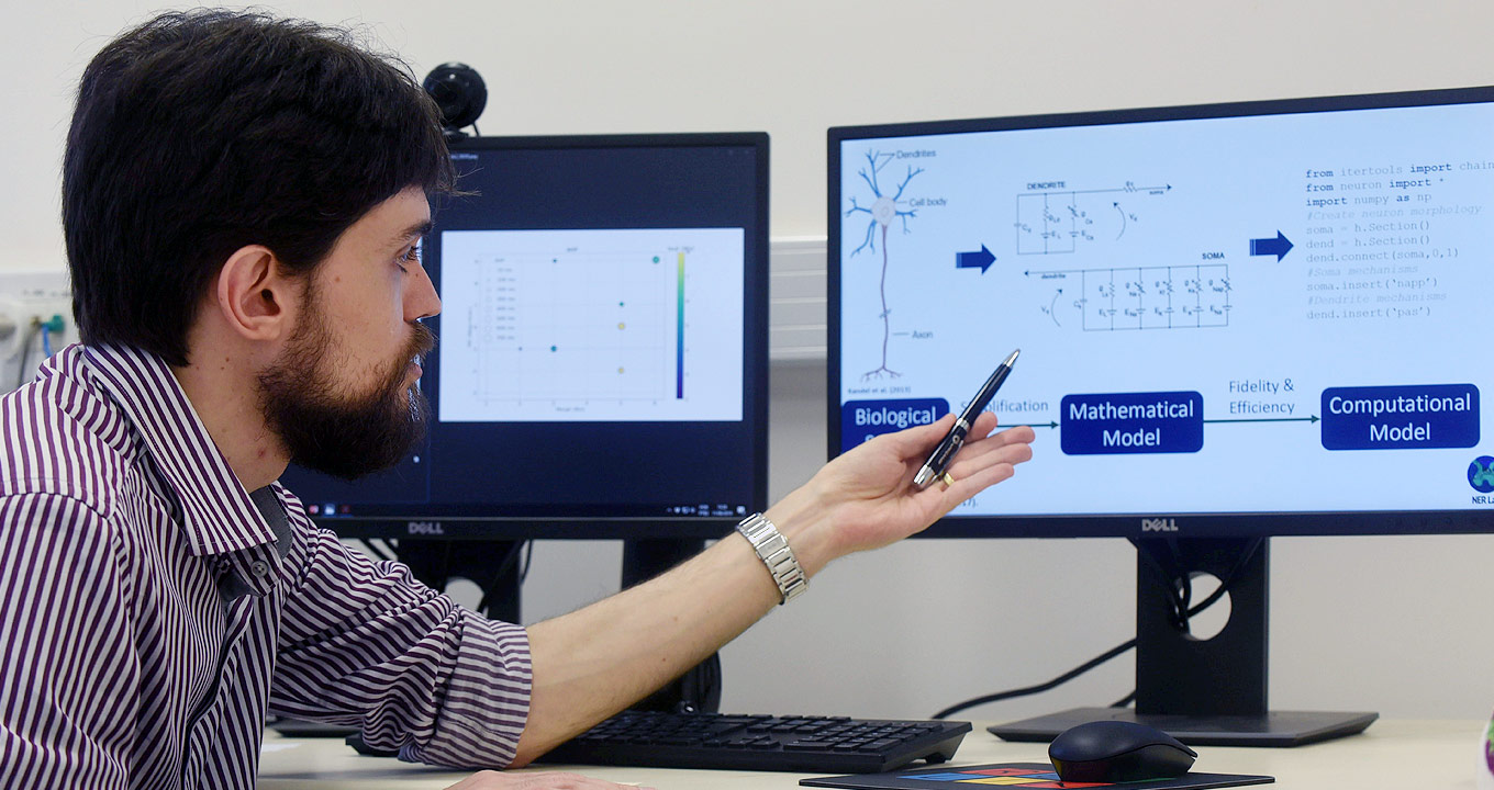 Audiodescrição: Em sala, imagem close-up e em perfil, homem, à esquerda na imagem, aponta com o braço esquerdo para monitor de lcd, à frente, esticando-o para frente e segurando uma caneta preta com a mão. O monitor, com cerca de vinte e duas polegadas, exibe imagem de neurônio, texto em inglês e um fluxograma. Ao lado desse monitor, há um outro, ambos sobre suposta mesa. O homem usa relógio no pulso esquerdo e camisa social com listras estreitas em vermelho e branco. Imagem 1 de 1.