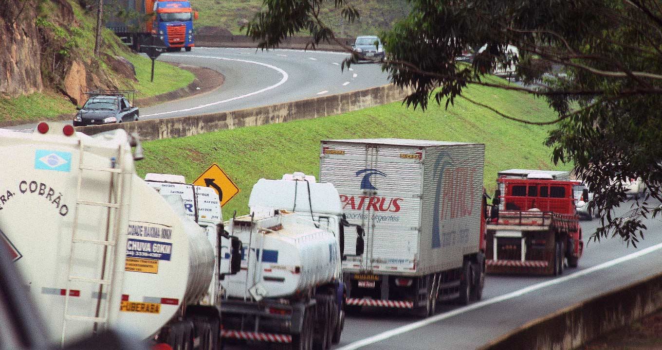 Audiodescrição: Imagem de trecho de uma rodovia com pista dupla, sendo que em primeiro plano, aparecem vários veículos de carga, como caminhões de carga, baú e pipa, um atrás do outro enfileirados, que trafegam da esquerda para a direita. Há um canteiro central gramado, com talude e com uma mureta de concreto, em desnível, que separa da pista que segue na direção oposta. O tempo está nublado. Imagem 1 de 1.