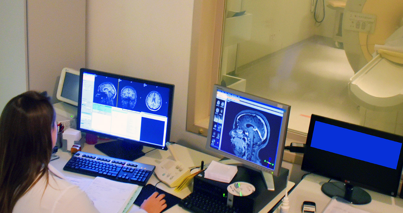 Neuroimagens no Hospital de Clínicas da Unicamp | Foto: Antoninho Perri Audiodescrição: em sala de exames com dois ambientes, imagem de costas e em perspectiva de uma mulher sentada em cadeira em cadeira, vestindo jaleco, tendo à frente uma bancada retangular de madeira sobre a qual estão quatro monitores de lcd. Dois deles mostram na tela imagens de ressonância magnética de cérebro humano. À frente da mesa há um grande vidro transparente quadrado, com cerca de um metro por um metro, que permite ver o outro ambiente, onde há um aparelho de ressonância magnética. Imagem 1 de 1.