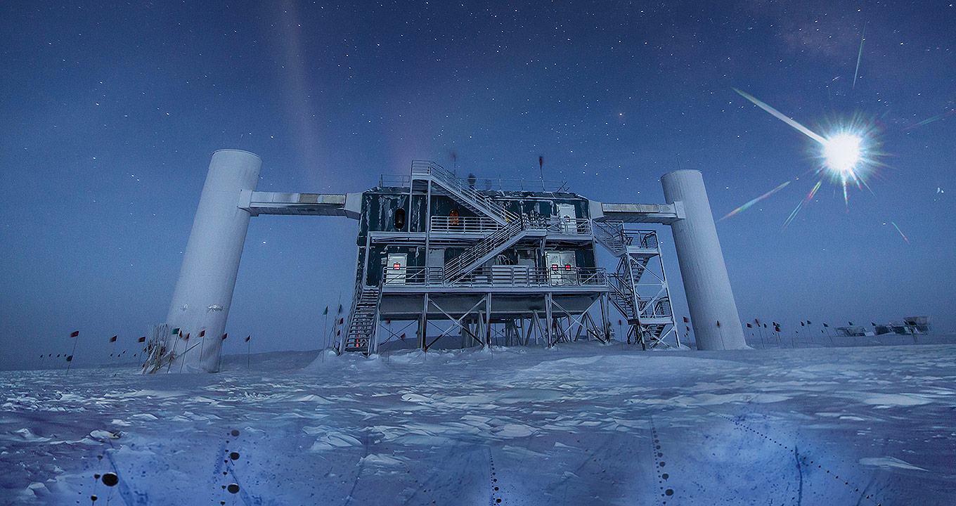 Observatório de Neutrinos IceCube | Reprodução | ceCube NSF Audiodescrição: Em área externa, imagem panorâmica e frontal da fachada de um observatório na Antártida, sendo que à esquerda e à direita há duas altas e largas colunas cilíndricas que estão interligadas á uma estrutura localizada entre elas, com dois andares e várias escadas. Esta estrutura é construída em metal e fica suspensa do chão, que está todo recoberto por neve ao redor do observatório. A imagem é escura, sendo que ao fundo aparece o céu repleto de estrelas e o sol. Imagem 1 de 1.