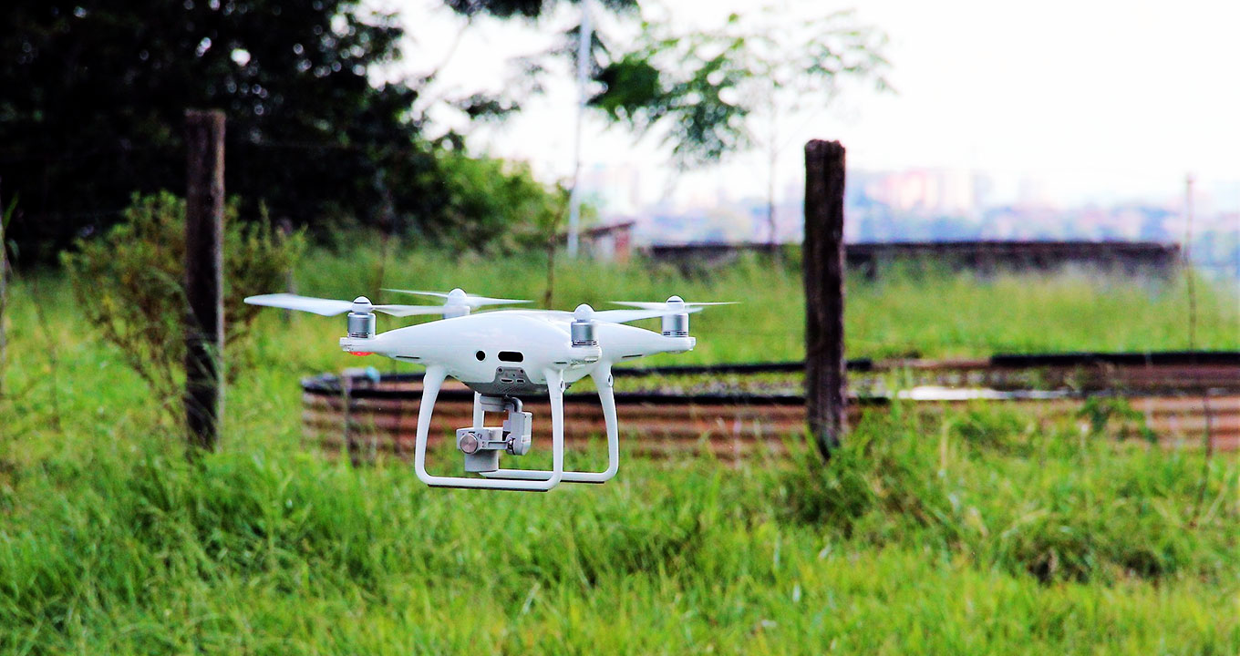 Audiodescrição: Drone de médio porte, branco, sobrevoa a baixa altura ampla área ao ar livre com vegetação rasteira. Ao fundo na imagem, há dois caibros de madeira afixados no chão que foram cerca de arame farpado. Imagem 1 de 1.