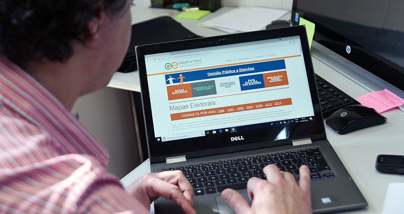 Audiodescrição: Em sala de trabalho, imagem close-up e em perspectiva, homem sentado em suposta cadeira, à esquerda na imagem, digita com as mãos em notebook que encontra-se sobre uma mesa de madeira à frente dele, sendo que aparecem apenas partes do rosto, do ombro, das costas e das mãos do homem. Sobre a mesa há também um teclado, um mouse e um celular. Na tela do notebook se lê em destaque Observatório das Eleições, Mapas Eleitorais. Imagem 1 de 1.