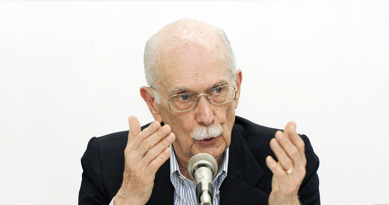 O professor, crítico e ensaísta Antonio Candido durante encontro que celebrou, em 2006, o 30º aniversário do Instituto de Estudos da Linguagem da Unicamp, unidade fundada e dirigida por ele | Foto: Antoninho Perri Audiodescrição: Em área interna, imagem frontal e de busto, homem ao centro na imagem fala em microfone afixado em pedestal de mesa, sendo que ele gesticula com os braços, mantendo os cotovelos apoiados sobre uma suposta mesa, os braços paralelos e com as mãos abertas. Ele usa óculos e veste terno preto sobre camisa social. Imagem 1 de 1.