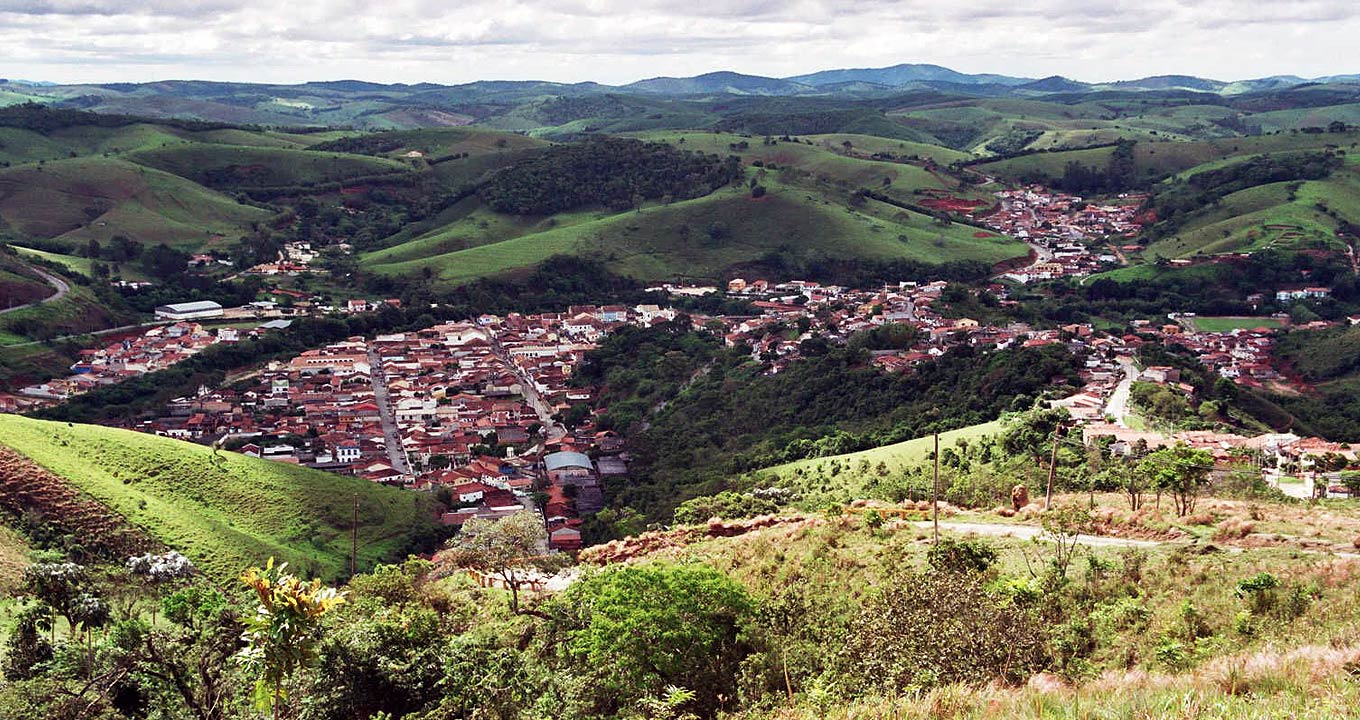 Audiodescrição: Em imagem panorâmica, vista aérea de uma cidade pequena, com várias casas agrupadas em bairros, sendo que entre esses núcleos há imensas áreas verdes e alguns morros cobertos por vegetação, que se estendem para todo o fundo da foto, fazendo limite com o céu nublado e com nuvens. Imagem 1 de 1.