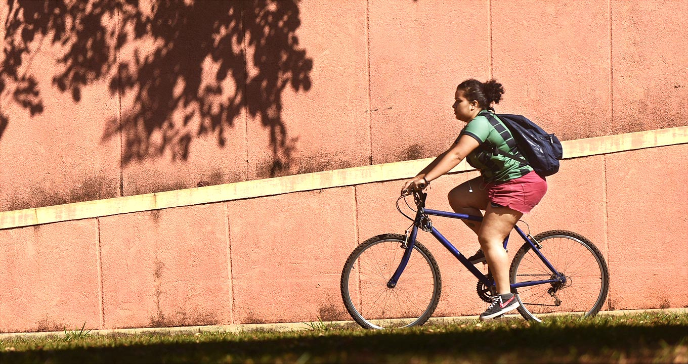 Audiodescrição: Em área externa bastante ensolarada, imagem em perfil e de corpo inteiro, mulher anda de bicicleta, ao centro na imagem, da direita para a esquerda. Ela circula rente a uma grande parede chapiscada, pintada em tom salmão, e que possui ranhuras verticais de metro em metro. Nela também há uma faixa de cimento com cerca de quinze centímetros de largura e que cruza toda a imagem na diagonal. A mulher usa bermuda curta vermelha, camiseta verde e leva uma mochila preta nas costas. Imagem 1 de 1.