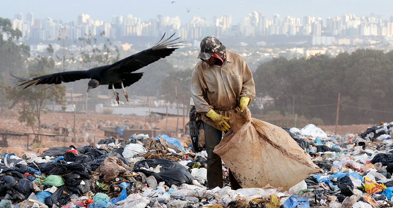 Catador de lixo em aterro sanitário na cidade de São Paulo | Foto: Flickr - Leopoldo Silva | Agência Senado Audiodescrição: Em área externa, imagem frontal e de corpo inteiro, homem em pé, ao centro na imagem, em um lixão, com resíduos espalhados em seu entorno. Ele segura um grande saco plástico com a mão esquerda, enquanto deposita material dentro do saco. Ele mantém a cabeça baixa e olhar voltado à esquerda. Ao lado dele, à esquerda, um urubu sobrevoa a área, com as asas abertas. A montagem de lixo está em lugar alto, de forma que aparecem em imagem panorâmica, ao fundo, inúmeros prédios e casas. Imagem 1 de 1
