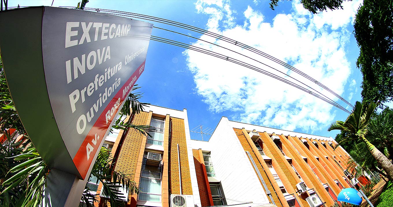 Fachada da Agência de Inovação da Unicamp | Foto:Thomaz Marostegan Audiodescrição: Em área externa, imagem panorâmica de baixo para cima, fachada de edificação com três andares e frente de tijolinhos e janelas de vidro transparente instaladas na vertical, formando colunas. À esquerda, placa metálica de cerca de sessenta centímetros de largura por oitenta de extensão, onde se lê em destaque Extecamp Inova, Prefeitura Universitária - Ouvidoria. Ao fundo do prédio, céu azul com nuvens. Fios da rede elétrica cruzam o céu na horizontal. À direita, algumas árvores. Imagem 1 de 1