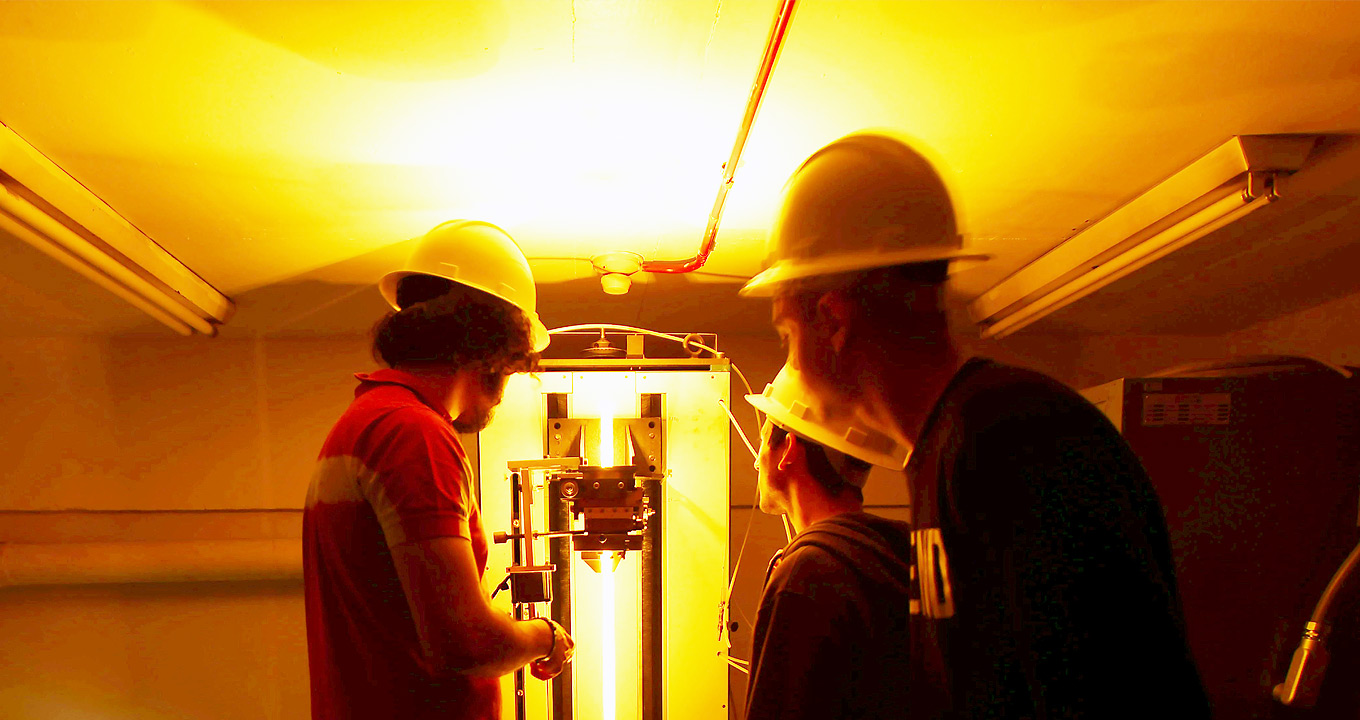 Foto: Antonio Scarpinetti Audiodescrição: Em laboratório com luzes amarelas e laranjas refletindo em todo ambiente, imagem em plano médio e de costas, três homens em pé, agrupados, sendo dois à direita e um à esquerda. Eles observam o homem à esquerda manusear um equipamento que emite luz na vertical, de cima para baixo, que se encontra à frente deles. Todos usam capacete de obra. O teto do local e baixo e tem luzes florescente que estão desligadas. Imagem 1 de 1.
