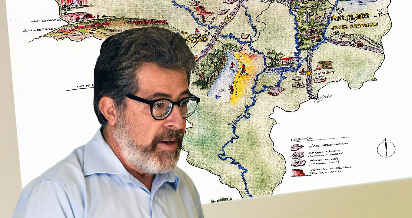 O professor Pedro Wagner Gonçalves e o mapa ilustrativo do Geoparque Corumbataí | Foto: Antonio Scarpinetti Audiodescrição: Em área interna, imagem em perspectiva e de busto, homem em pé, ao centro na imagem e com olhar voltado para a direita, sendo que ele mantém os braços abaixados. Às costas dele há um mapa ilustrado com pinturas em lápis de cor, com detalhes de áreas de vegetação, em verde, e fluxo de rios, em azul. Ele usa óculos e veste camisa social em tom azul claro. Imagem 1 de 1.