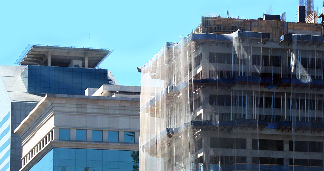 Audiodescrição: Em área ao ar livre, imagem panorâmica e em perspectiva, parte superior de um prédio ainda em construção, à direita na imagem, com escoras de madeira e ferragens à mostra, e com telas de proteção feitas de nylon em todo o entorno. Logo atrás desse prédio, há um outro, já finalizado, onde se destacam modernas janelas de vidro nas laterais e um heliponto na parte superior. O dia está bastante ensolarado, com céu azulado aparecendo ao fundo e à esquerda na imagem. Imagem 1 de 1.