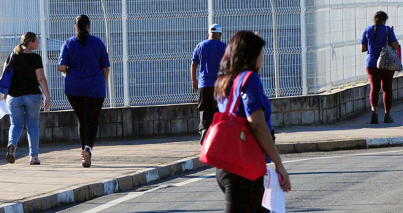 Audiodescrição: Em área externa, imagem de corpo inteiro e de costas, cinco pessoas caminham em fila, espaçada, sobre uma calçada de cimento, da esquerda para a direita na imagem. Elas andam rentes a uma grade de branca, afixada em uma mureta com cerca de quarenta centímetros, construída com blocos de concreto. A calçada tem uma curva para a direita. Também aparece parte da rua asfaltada, por onde uma mulher circula, ao centro na imagem. O dia está ensolarado. Imagem 1 de 1.