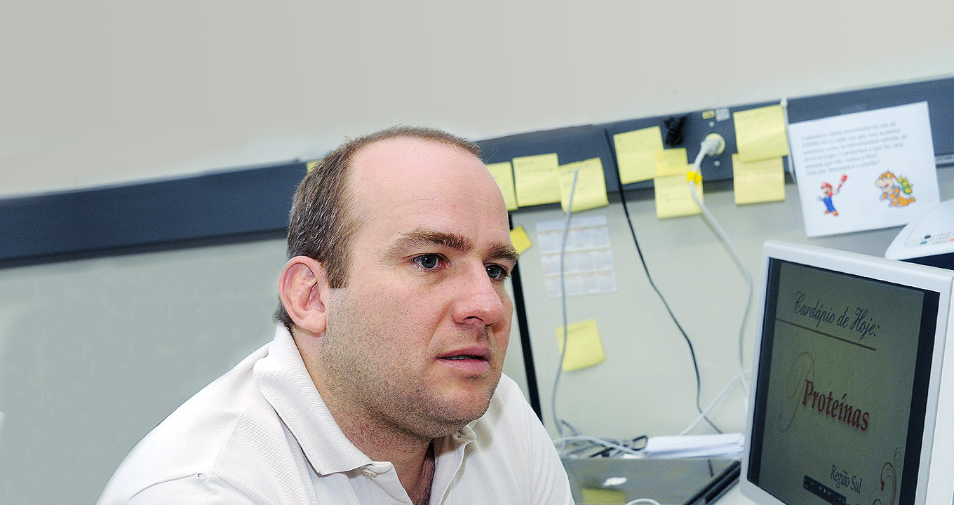Eduardo Galembeck, professor do Instituto de Biologia e responsável pelo desenvolvimento do software | Foto: Antonio Scarpinetti Audiodescrição: Em sala de trabalho, imagem em perspectiva e close-up, homem sentado em suposta cadeira, com olhar voltado para a direita, fala mantendo os braços baixados, sendo que à frente dele há um monitor de lcd de cerca de vinte e duas polegadas apoiado sobre uma mesa, e onde se lê em destaque cardápio de hoje: proteínas. Ao lado dele, ao fundo da imagem, há um parede com dezenas de pequenos papéis de anotação de cor amarelo afixados. Ele veste camisa polo branca. Imagem 1 de 1.