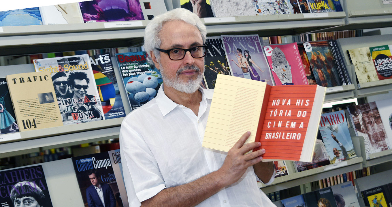 Fernão Pessoa Ramos, professor do Instituto de Artes da Unicamp e um dos organizadores do livro “Nova história do cinema brasileiro” | Foto: Antonio Scarpinetti Audiodescrição: Em área interna, imagem em perspectiva e de busto, homem em pé, ao centro e com olhar para as lentes da câmara fotográfica. Ele segura com as mãos um livro aberto na primeira página, erguendo-o na altura do ombro, à direita, e trazendo-o junto ao corpo. O livro tem formato A4 e cerca de três centímetros de espessura. Na página aberta se lê Nova História do Cinema Brasileiro. Às costas dele, há um tipo de estante de ferro com três prateleira repleta de revistas com capa de arte. Imagem 1 de 1