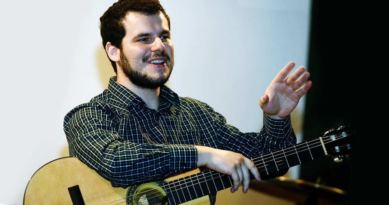 O violonista Victor Polo, autor da pesquisa | Foto: Antonio Scarpinetti Audiodescrição: Em área interna, imagem em perspectiva e em plano médio, homem sentado, com olhar voltado para a direita, sorri tendo apoiado no colo um violão. Ele segura o braço do violão com a mão direita e mantém o esquerdo levantado e projetado para frente, com a palma da mão aberta. Ele veste camisa com riscos formando xadrez em tom claro sobre fundo preto. Imagem 1 de 1.