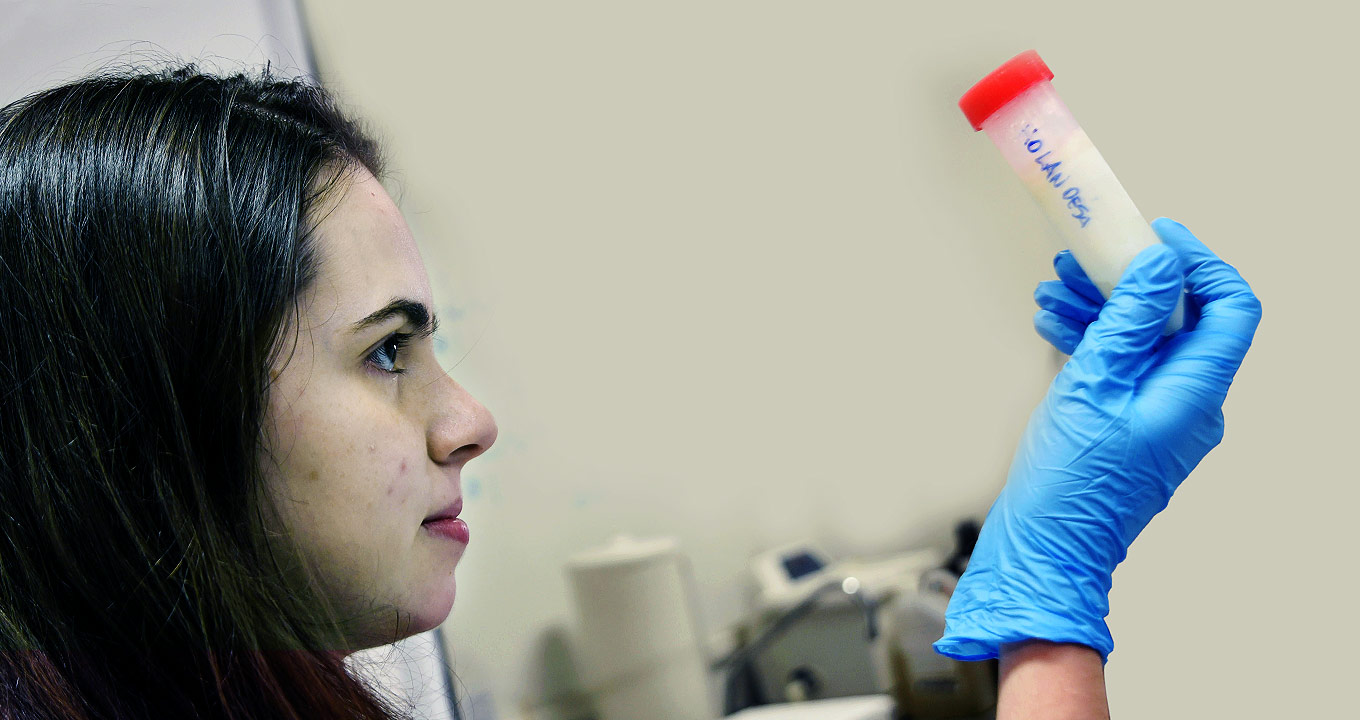 A pesquisadora Jeany Delafiori analisa amostra de leite no Laboratório Innovare de Biomarcadores | Foto: Antonio Scarpinetti Audiodescrição: Em área interna, imagem em perfil e close-up, mulher com o olhar voltado para a direita mantém o braço direito erguido na altura dos olhos, sendo que ela segura entre o dedos um tubo plástico com cerca de quinze centímetros de comprimento por cinco de diâmetro, com uma tampa vermelha. O tubo contém líquido branco e em sua lateral está escrito com caneta holan oesa. Ela usa uma luva azul de silicone. Ao fundo há uma parede bege e aparece parte de alguns equipamentos. Imagem 1 de 1.