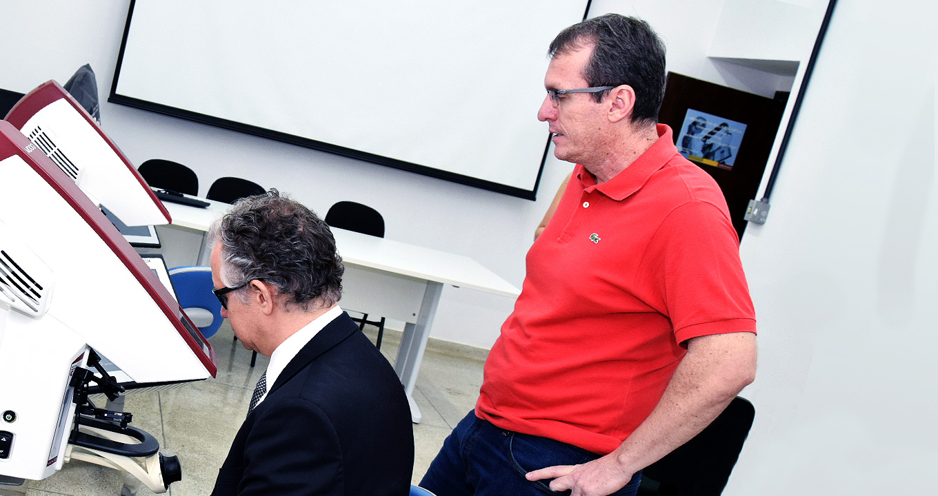 Audiodescrição: Em sala de aula, imagem em perfil e de busto, homem sentado em cadeira, ao centro e com olhar voltado para a esquerda, manuseia equipamento que se encontra à frente dele sobre uma mesa, usando óculos especial. O equipamento é um tipo de simulador usado na área de odontologia. Atrás dele, um outro homem em pé o observa. Há outro equipamento idêntico à direita. Ao fundo da imagem há uma tela de projeção afixada na parede e uma mesa retangular branca de madeira com três cadeiras. Imagem 1 de 1.