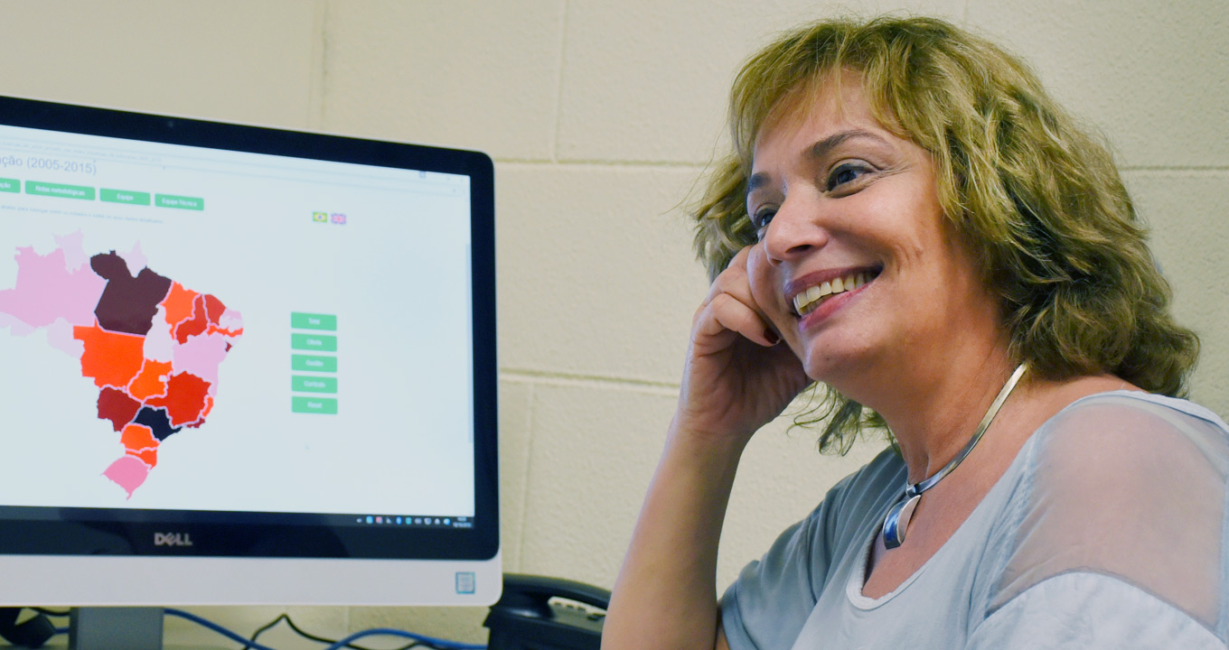 A professora Theresa Adrião, coordenadora da pesquisa | Foto: Antoninho Perri Audiodescrição: Em sala, imagem em perfil e de busto, mulher sentada em cadeira sorri, sendo que ela mantém o cotovelo direito apoiado sobre suposta mesa à frente dela, com a mão direita encostada na face direita do rosto. Sobre a mesa, à esquerda, há um monitor de lcd com cerca de trinta polegadas que exibe mapa do Brasil, com os estados que o compõem demarcadas com cores como laranja, rosa, preto e marrom. Ela usa um colar prateado e uma blusa branca. Ao fundo, parede de blocos de concreto. Imagem 1 de 1.