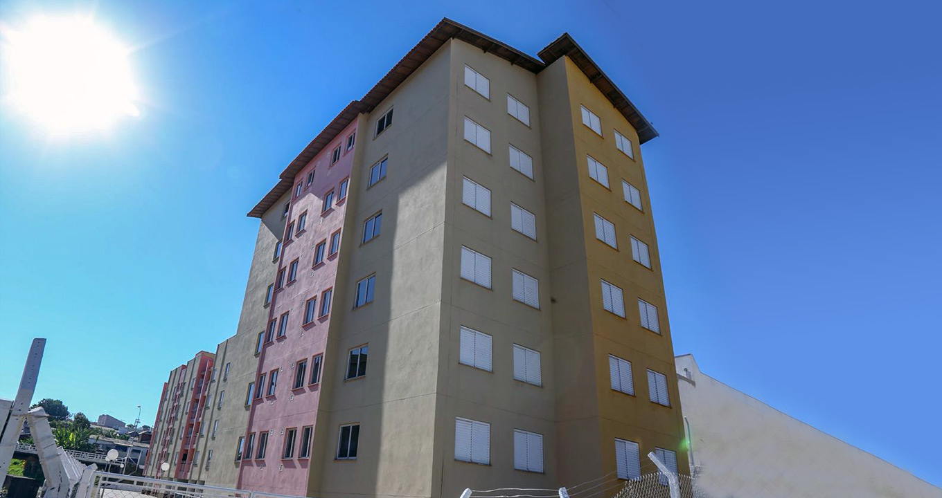 Novo conjunto habitacional da CDHU em Marília, em foto de setembro deste ano | Foto: Amanda B. Donatiello/CDHU Fotos. Em área externa, imagem panorâmica e de baixo para cima de um conjunto habitacional, sendo que ao centro há um prédio de seis andares, simples, com janelas quadradas e padronizadas em toda a fachada e na lateral à esquerda. À esquerda na imagem, fachada de outro conjunto habitacional. Ao fundo de toda a imagem, céu bastante azulado com o sol brilhando à esquerda. Imagem 1 de 1.