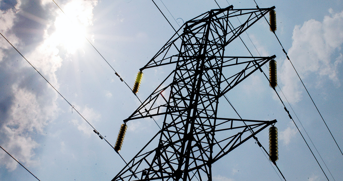 Audiodescrição: Em área externa, imagem de baixo para cima e em perspectiva, torre de transmissão de energia elétrica, com mais de vinte metros de altura e vários cabos paralelos afixados nela ela, à direita e à esquerda, que cruzam a imagem, paralelos e na diagonal. O sol brilha entre nuvens, à esquerda na imagem, sendo que há outras nuvens esparsas no céu azul. Imagem 1 de 1.