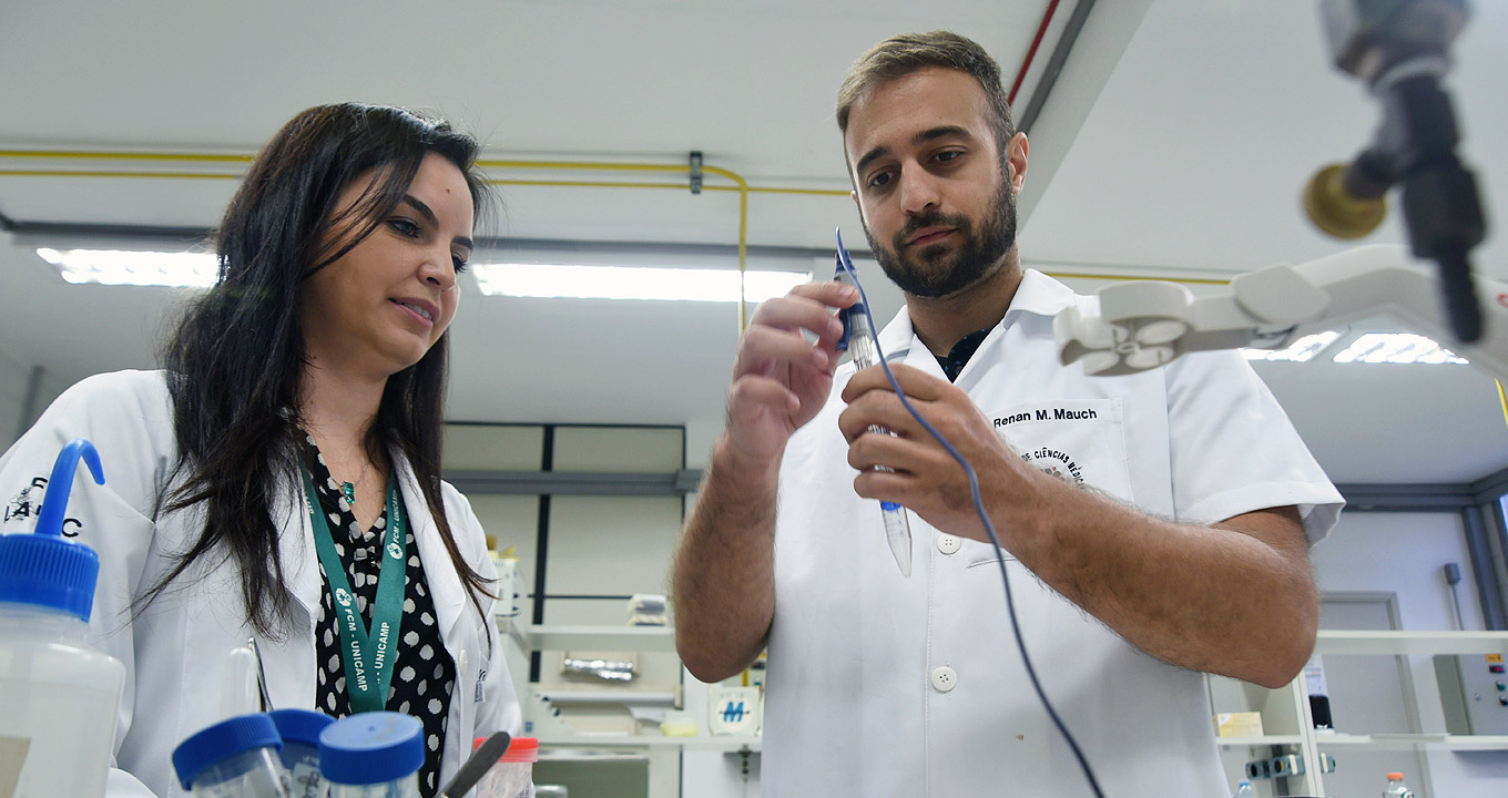 A fisioterapeuta Carla Cristina Souza Gomez e o biomédico Renan Marrichi Mauch em laboratório na FCM | Foto: Antonio Scarpinetti Audiodescrição: em laboratório, imagem frontal e de busto, mulher, à esquerda, e homem em pé, um ao lado do outro, ao centro na imagem. Ele manipula com as mãos um equipamento composto por um tipo de tubo de ensaio de vidro que tem um longo e fino cabo afixado ao bocal do tubo de ensaio, enquanto a mulher o observa. À frente deles, há uma bancada com outros objetos encima, como potes plásticos. Ele veste jaleco branco de mangas curtas com nome no bolso, e ela jaleco branco de mangas compridas. Imagem 1 de 1