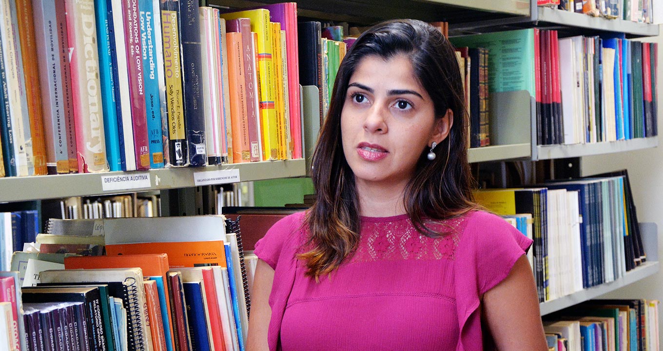 A fonoaudióloga Núbia Garcia Vianna, autora da pesquisa | Foto: Mário Moreira Audiodescrição: em uma biblioteca, imagem em perspectiva e de busto, mulher em pé com olhar voltado para a esquerda, fala mantendo os braços baixados. Rente à ela, à esquerda na imagem, uma extensa estante de ferro com três prateleiras repletas de livros. Ele veste blusa sem mangas em tom rosa. Imagem 1 de 1.