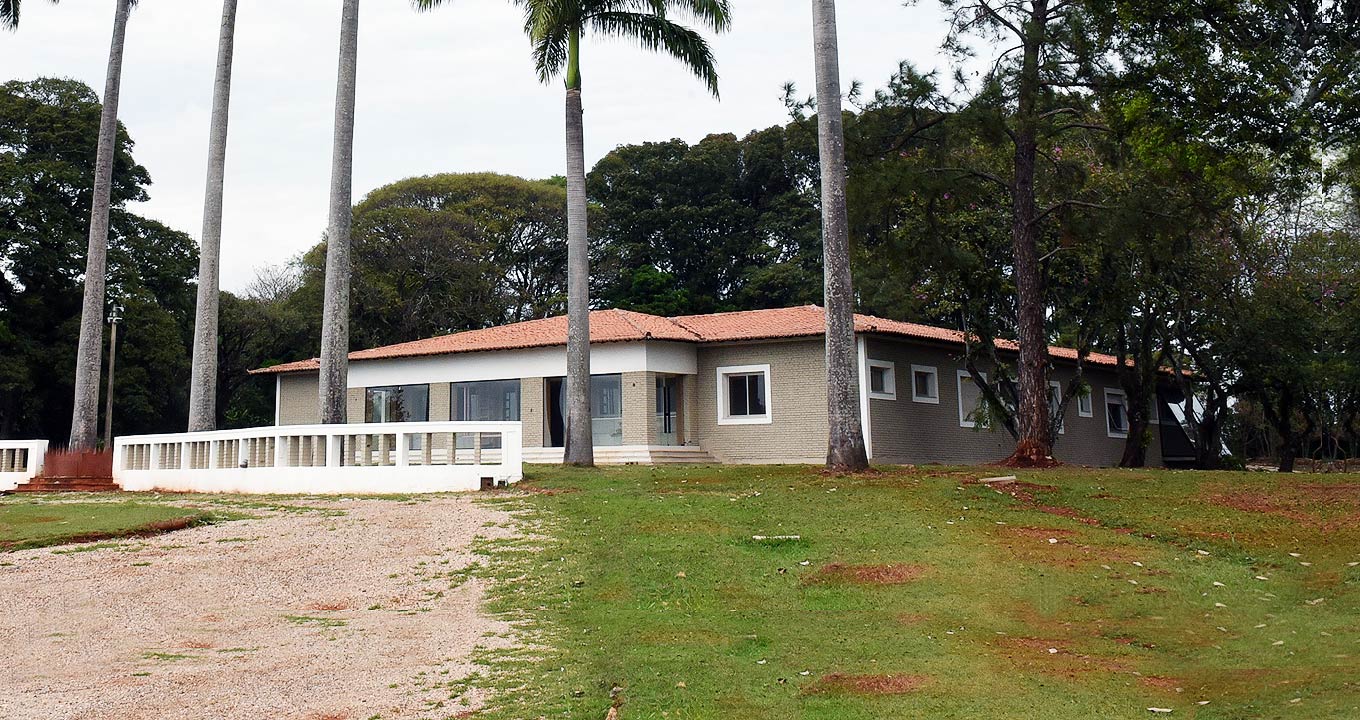 Audiodescrição: Em área externa, imagem panorâmica e em perspectiva, ampla casa de campo com janelas retangulares à frente e à direita na imagem. À frente da casa há oito altas palmeiras imperiais dispostas uma ao lado da outra, alinhadas na horizontal, com intervalos de três metros. No entorno da casa, ampla área gramada com caminho de areia para passagem de veículos. À frente há também uma extensa mureta com cerca de oitenta centímetros de altura. Ao fundo ampla vegetação com árvores altas. Imagem 1 de 1.