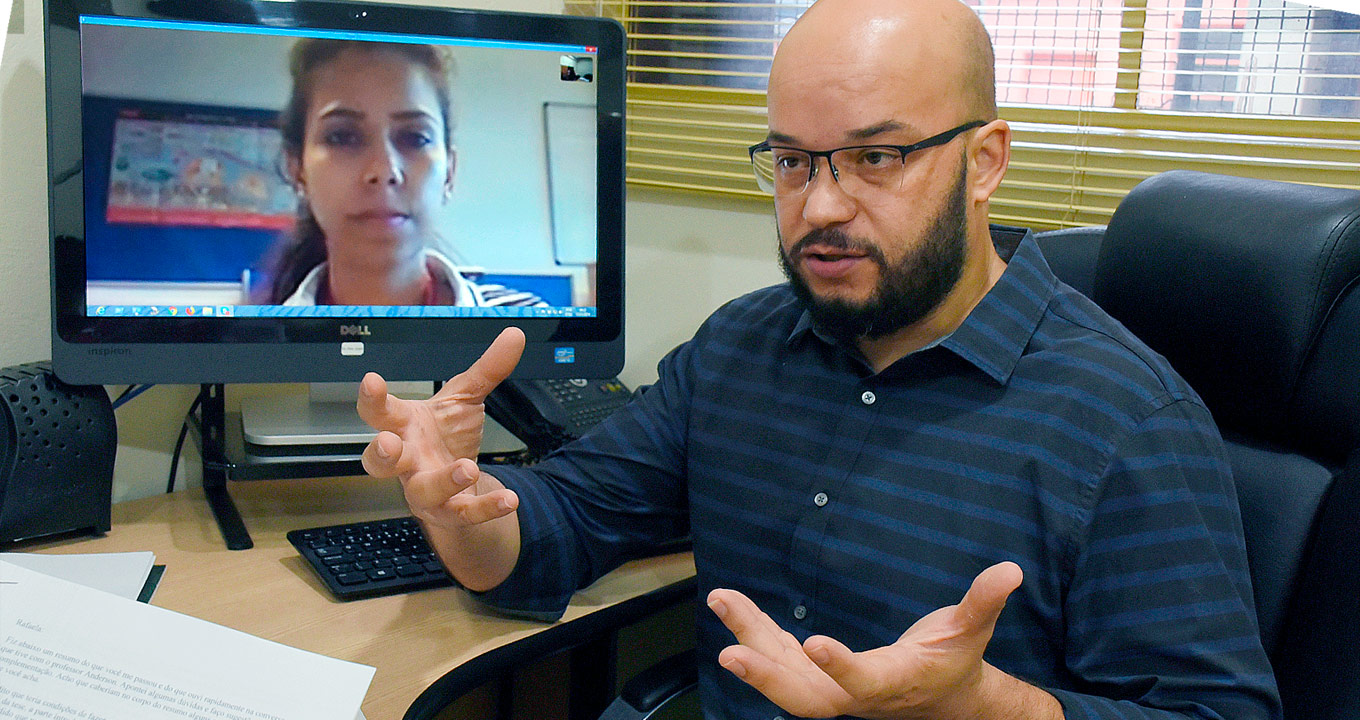 O professor Anderson de Souza Sant’Ana, orientador da pesquisa, e, na tela, Rafaela de Carvalho Baptista, autora | Foto: Antoninho Perri Audiodescrição: em sala de trabalho, imagem em perspectiva e de busto, homem sentado em cadeira, com olhar voltado para a esquerda, fala e gesticula com as mãos, mantendo os braços projetados para frente e palmas das mãos abertas, com os dedos separados. Logo à esquerda, junto a ele, sobre uma suposta mesa, um monitor de lcd com cerca de 32 polegadas que exibe imagem frontal e close-up do rosto de uma mulher. Às costas dele, à direita, janela com persianas verticais, e à frente, uma mesa. Imagem 1 de 1.
