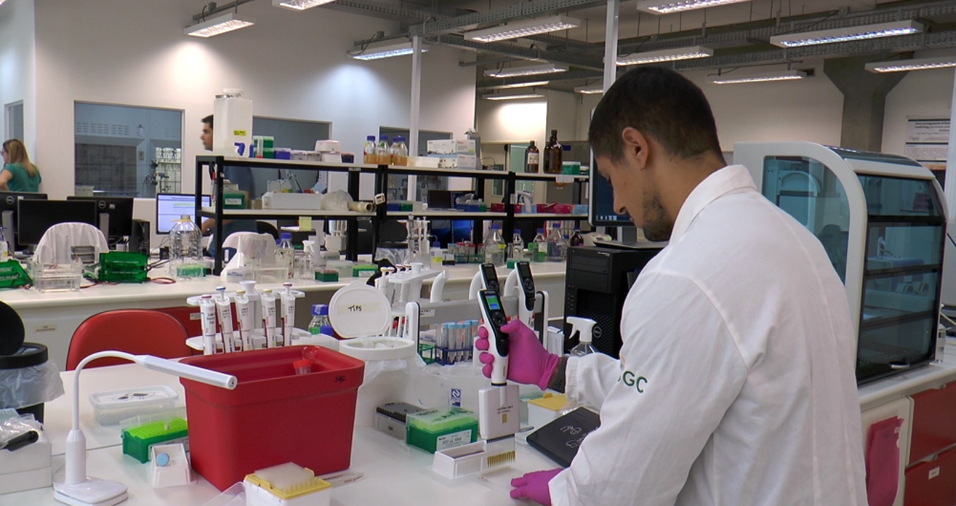 Centro de Química Medicinal (CQMED) desenvolve pesquisas em colaboração com Aché e Eurofarma e pretende fechar acordo com mais três farmacêuticas sediadas no país até o fim de 2019 | Foto: Daniel Antônio | Agência FAPESP Audiodescrição: em amplo laboratório de química, imagem em perspectiva, de busto e de costas, homem à direita na imagem e olhar voltado para a esquerda, manipula com a mão direita um tipo de controle remoto com painel digital. À frente dele há uma extensa bancada branca retangular de madeira, sobre a qual se encontram outros equipamentos usados no laboratório. À frente desta bancada há outras duas iguais. Há outras duas pessoas em pé no laboratório. Ele usa jaleco branco de mangas compridas. Imagem 1 de 1.