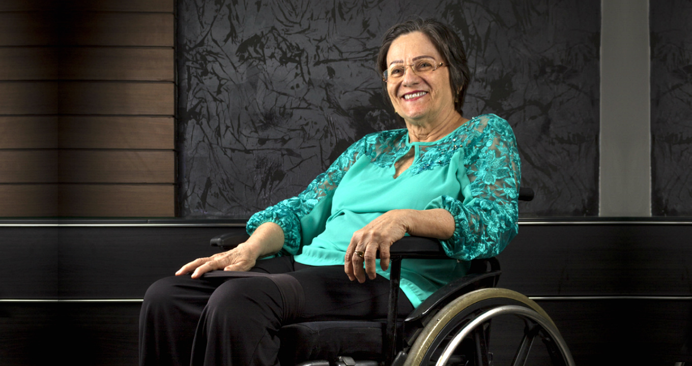 A farmacêutica Maria da Penha Maia Fernandes, que participa do evento “Futuro da Mulher no Mundo do Trabalho” | Foto: Chico Max Audiodescrição: em área interna, imagem em perspectiva e de corpo inteiro, mulher em cadeira de rodas, ao centro na imagem, sendo que ela sorri, com olhar voltado para frente, mantendo o braço esquerdo apoiado sobre o braço da cadeira, e a mão direita espalmada sobre a coxa direita. Ela usa óculos e veste calça preta e blusa verde de mangas compridas com detalhes bordados nos ombros e no peito. Ao fundo, às costas dela, parede preta com textura e painéis horizontais de madeira. Imagem 1 de 1.