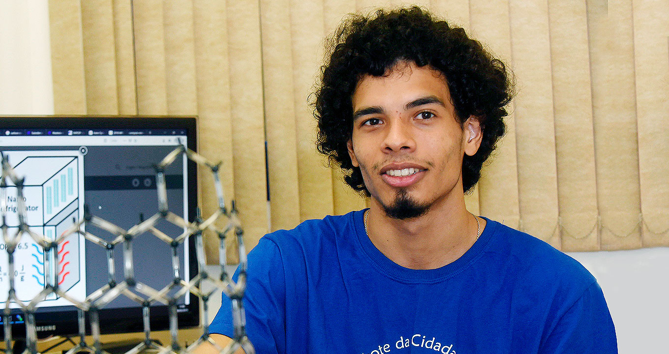 O estudante Tiago Cantuário, autor do trabalho | Foto: Antonio Scarpinetti Audiodescrição: em área interna, imagem frontal e de busto, jovem sorri, mantendo os braços baixados. À esquerda na imagem, junto a ele, há um monitor de lcd com cerca de 25 polegas que exibe ilustrações geométrica retangulares, e uma estrutura metálica construída com um tipo de arame com vários hexágonos unidos. Às costas dele, há persianas verticais que ocupam quase todo o fundo da imagem a partir da direita. Ele vete uma camiseta azul. Imagem 1 de 1.