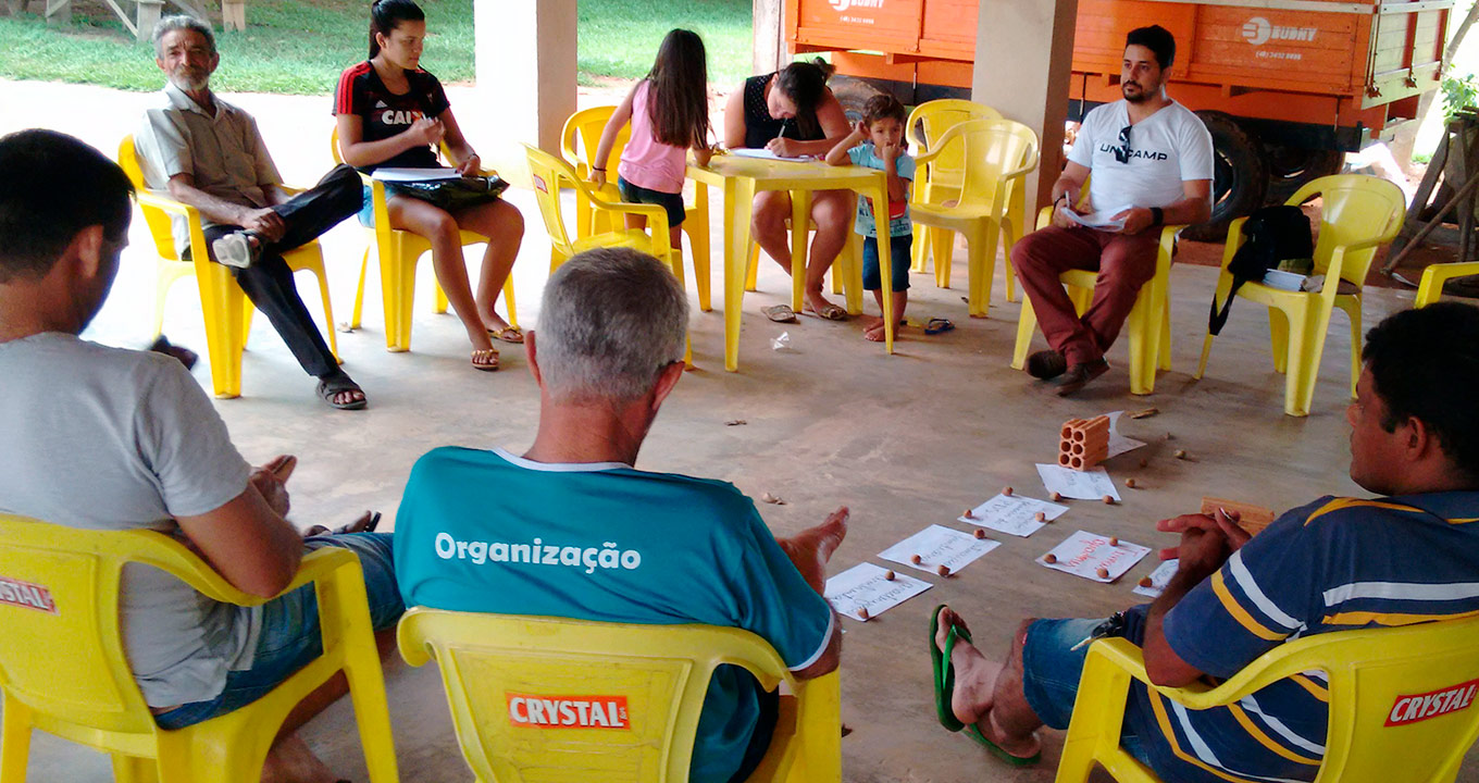 Trabalho de campo em um dos assentamentos: locais apresentam características distintas, desde o histórico e a forma de ocupação até os modelos de organização e produção | Foto: Divulgação Audiodescrição: em área coberta aberta, imagem frontal e de corpo inteiro, círculo formado por cerca de dez pessoas sentadas em cadeiras plásticas amarelas. Ao fundo, à direita, parte de uma carroceria de madeiras, e à esquerda, parte de uma área gramada e ensolarada. As pessoas usam roupas informais, como camiseta e chinelo, e algumas seguram caneta e folhas de papel. Ao centro da roda, há algumas folhas de papel sulfite no chão. Uma mulher escreve em folha de papel sobre uma mesa plástica. Imagem 1 de 1.
