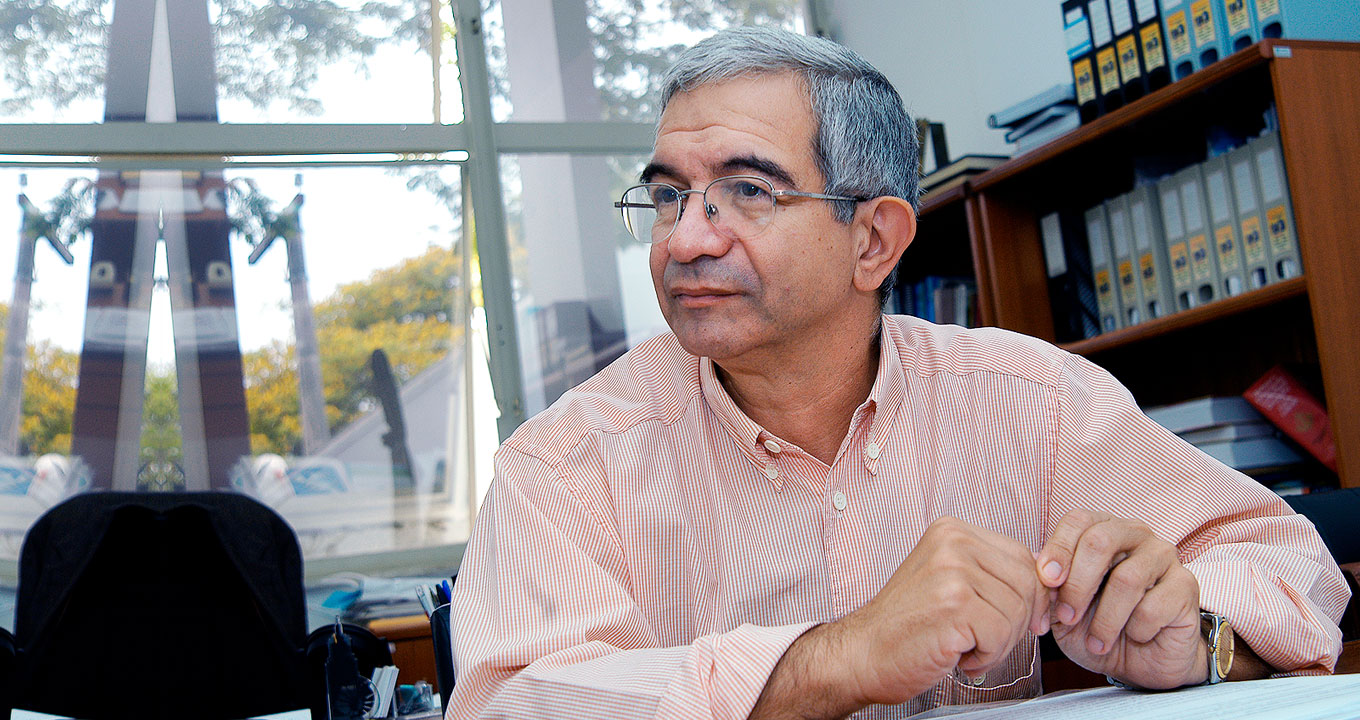 O professor Fernando Costa, único brasileiro entre os autores | Foto: Antoninho Perri Audiodescrição: em área interna, imagem de busto e frontal, homem sentado em cadeira, à direita na imagem, com rosto virado para a esquerda, sendo que mantém as mãos encostadas pela pontas de alguns dedos, com braços levemente projetados para frente. Ele usa óculos e veste camisa social de mangas compridas, com finas listras verticais em vermelho e branco. Às costas dele, à esquerda, estante de madeira com livros e pastas nas prateleiras. Ao fundo, uma janela retangular de vidro transparente. Imagem 1 de 1.