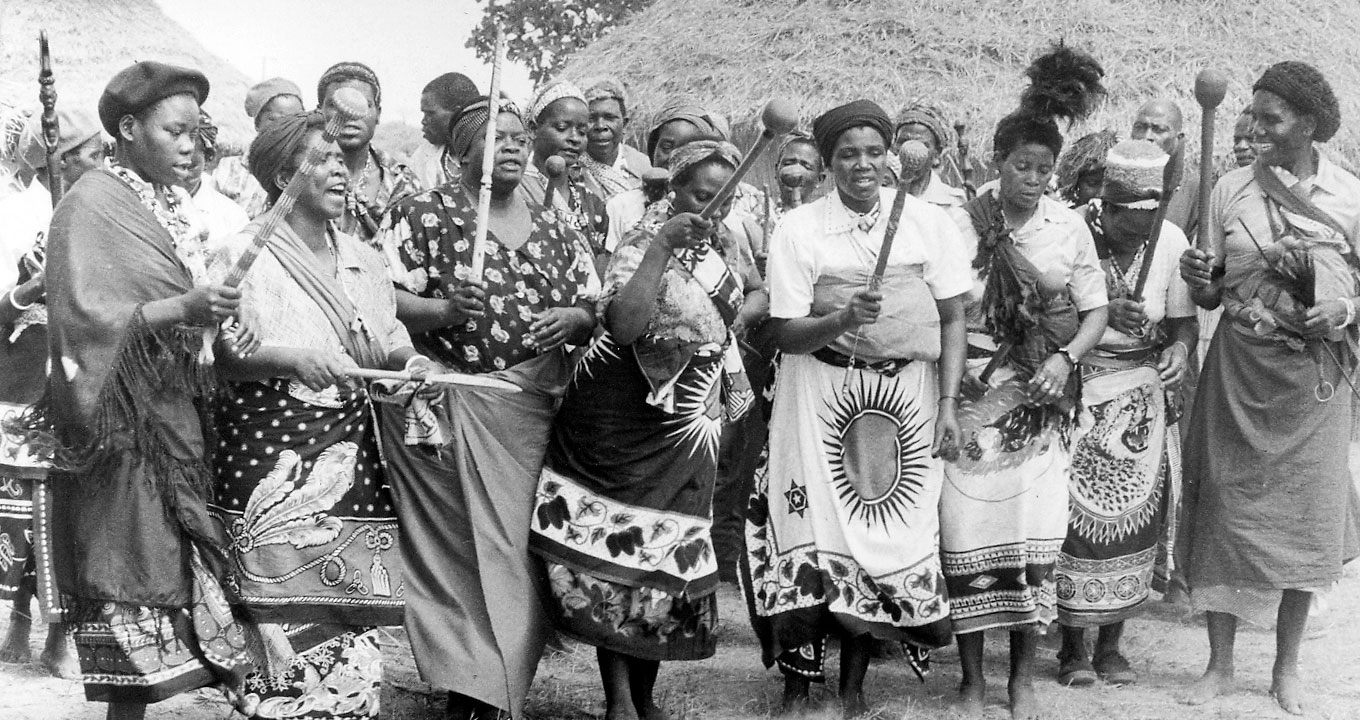 Grupos de tinyanga em Chaimite | Imagem: Reprodução Audiodescrição: Em área externa, imagem frontal e de corpo inteiro, cerca de vinte mulheres em pé, agrupadas horizontalmente, uma próxima à outra, sendo que todas elas são negras e usam um tipo de bandana africana e seguram com a mão direita um tipo de bastão típico da aldeia, com cerca de sessenta centímetros de comprimento e com uma bola na ponta. Às costas delas aparece parte do telhado de uma habitação feito de palha. A Imagem é em preto e branco e antiga. Imagem 1 de 1.
