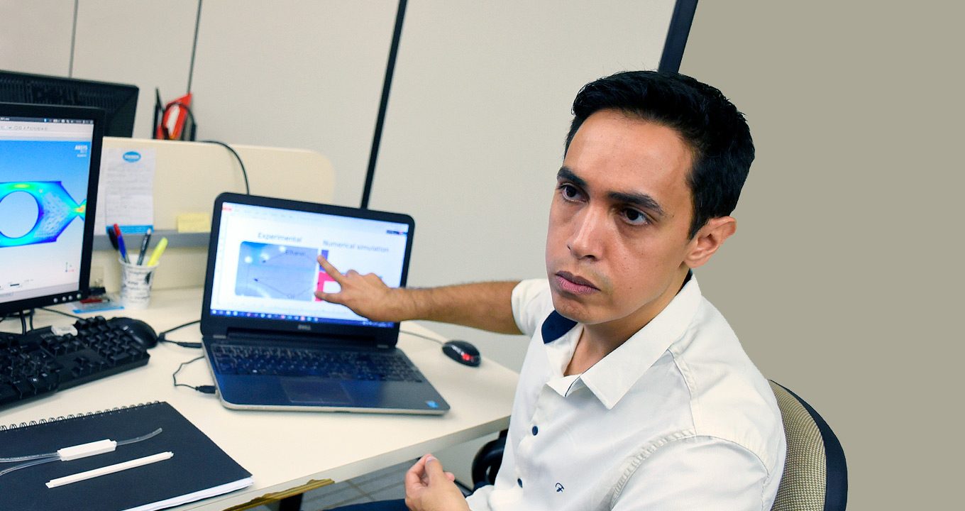 O engenheiro químico Harrson Santana, autor do trabalho: desafio da pesquisa foi ampliar o tamanho do dispositivo, sem que este perdesse eficiência | Foto: Antoninho Perri Audiodescrição: em sala de trabalho, imagem em perspectiva, de busto e de cima para baixo, homem sentado em cadeira com o olhar voltado para a esquerda da imagem, aponta com dois dedos da mão direita, esticando o braço, para tela de notebook que encontra-se ao lado dele, sobre uma mesa de madeira retangular. O braço esquerdo permanece apoiado sobre suas pernas. Na mesa há ainda um teclado de computador, um monitor de lcd e duas peças semelhantes e tiras de plástico, sobre a capa de um caderno. Imagem 1 de 1