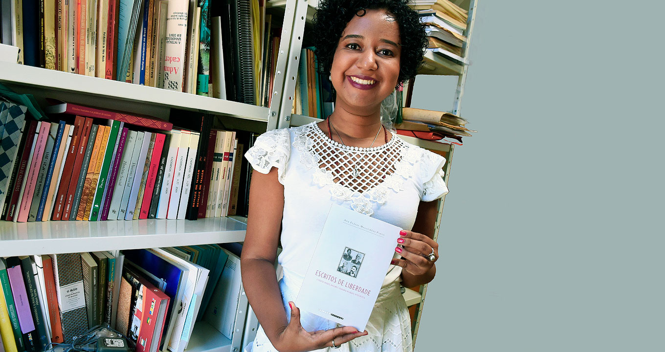 A historiadora Ana Flávia Magalhães Pinto, autora do livro “Escritos da Liberdade: Literatos negros, racismo e cidadania no Brasil oitocentista” | Foto: Antoninho Perri Audiodescrição: em uma sala, imagem frontal e de busto, mulher em pé, ao centro da imagem, sorri e segura com as duas mãos um livro de capa branca com quatro pequenas imagens. Às costas dela, à esquerda, uma estante branca de ferro, com três prateleiras repletas de livros. Ela usa uma blusa branca de mangas curtas. Imagem 1 de 1.