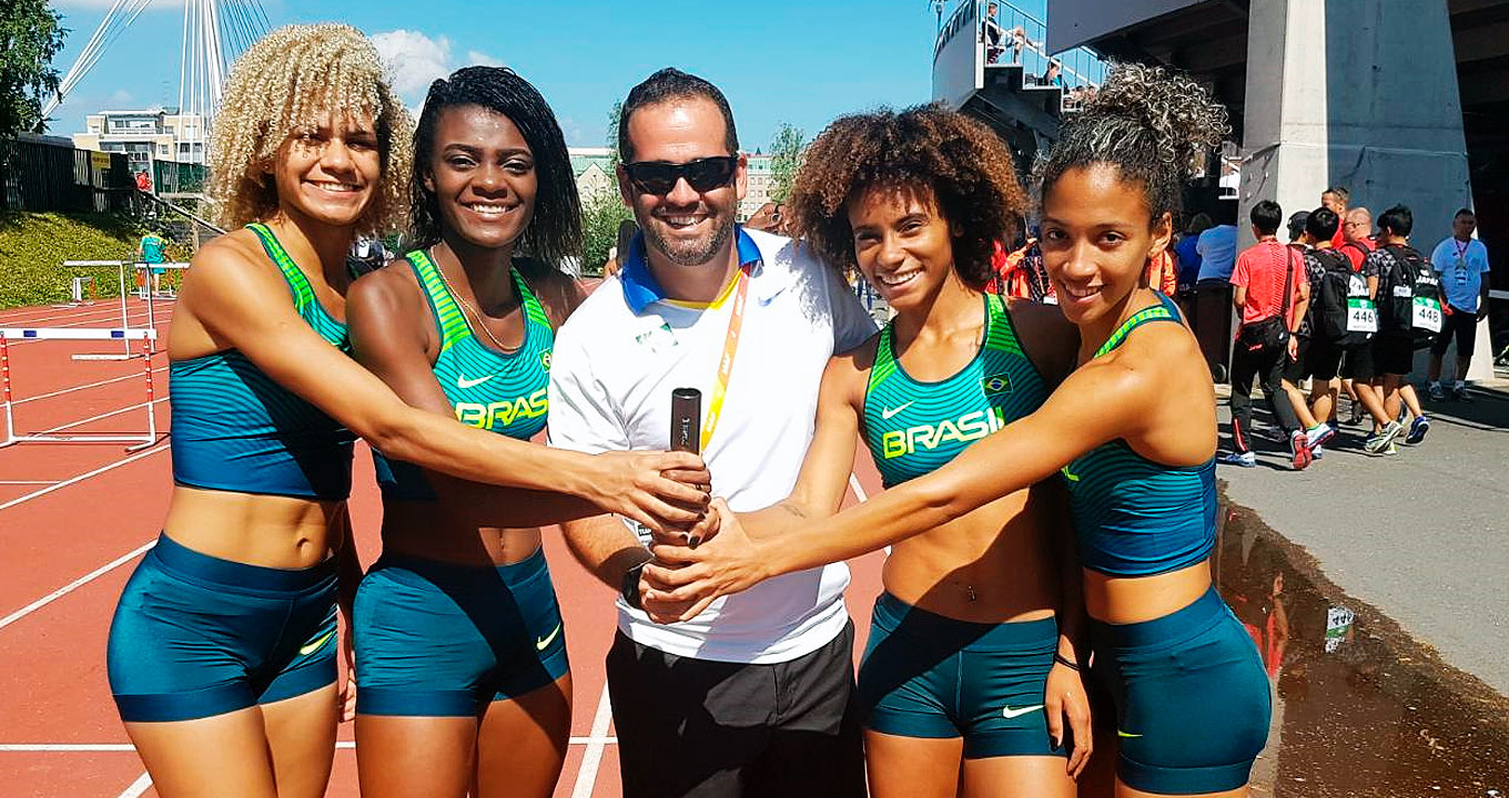 Audiodescrição: em ampla pista de atletismo, imagem frontal e em plano médio, quatro mulheres e um homem, ao centro, todos em pé, sorrindo, um ao lado do outro. O homem segura com a mão, projetando o braço para frente, um basta preto usado em corrida de revezamento, enquanto as mulheres esticam o braço em direção ao bastão e o colocam uma das mãos no bastão. Elas vestem traje de corrida, com bustier de lycra azul e verde, onde se Brasil em letras brancas, e short de lycra. O dia é ensolarado. Imagem 1 de 1.