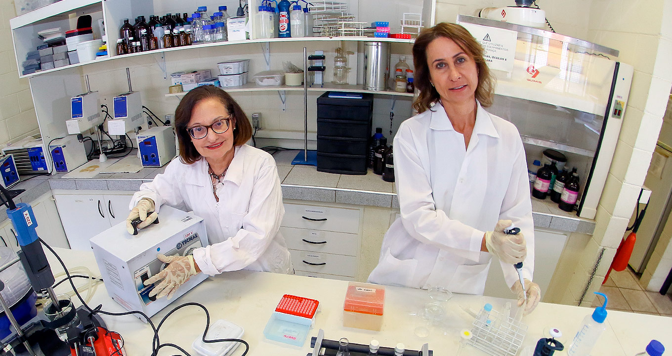 Audiodescrição: em laboratório farmacêutico, imagem frontal e em plano médio, duas mulheres em pé, uma ao lado da outra, sendo que elas mantêm o olhar voltado para as lentes da câmara fotográfica. A mulher à direita segura um tipo de dosador com a mão direita e um tubo de ensaio com a esquerda, enquanto a da esquerda opera um equipamento que está sobre um extenso balcão retangular de madeira à frente delas. Às costas, uma estante com material de laboratório. Elas usam luvas e jalecos brancos. Imagem 1 de 1.