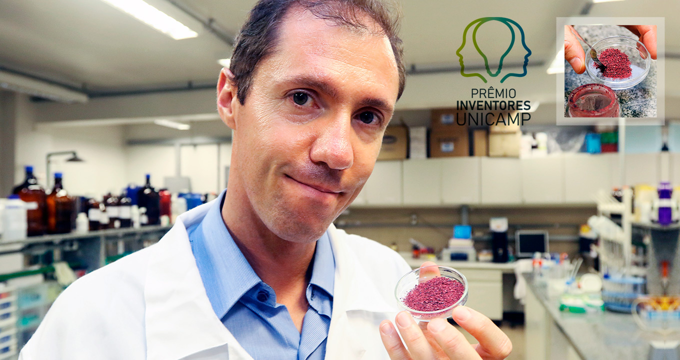 Audiodescrição: em laboratório de pesquisa, imagem frontal e de busto, homem ao centro da imagem e olhar voltado para a lente da câmera fotográfica, segura com as pontas dos dedos da mão esquerda um recipiente cilíndrico de vidro ou acrílico transparente, com cerca de oito centímetros de diâmetro por um centímetro de altura contendo farelo de cor roxa. Ao fundo há armários de madeira com portas e, nas laterais, bancadas com vários equipamentos e materiais de laboratório. Ele usa jaleco branco. Imagem 1 de 1