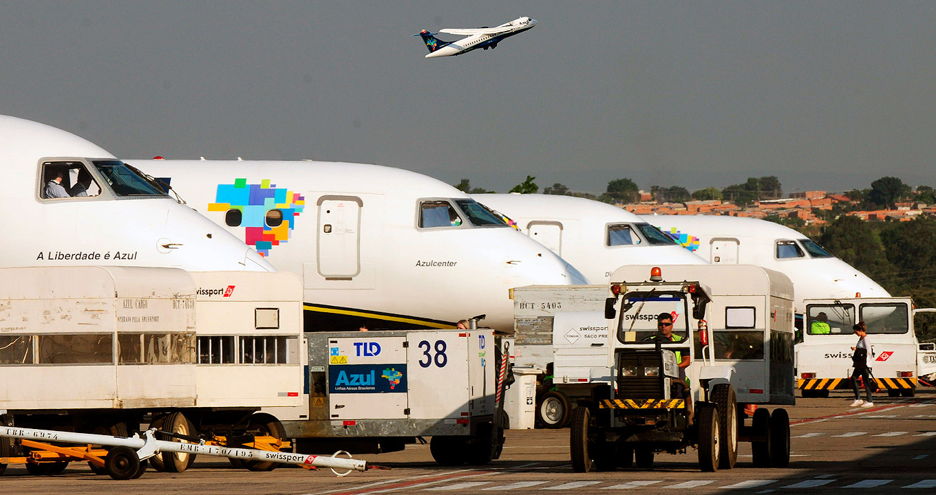 Aviões no Aeroporto de Viracopos, em Campinas | Foto: Antonio Scarpinetti Audiodescrição: no pátio de um aeroporto, imagem em perspectiva, 4 aeronaves de grande porte, à esquerda na imagem, estacionadas uma ao lado da outra, com proas voltadas para a direita. A foto mostra a parte frontal das cabines das aeronaves, todas branca, sendo que na primeira delas, aparecem duas pessoas na janelinha. À frente dos aviões, há veículos de apoio às aeronaves, e ao fundo na imagem, várias casas edificadas com tijolo, sem acabamento, em meio à vegetação. Um avião decola ao fundo. Imagem 1 de 1