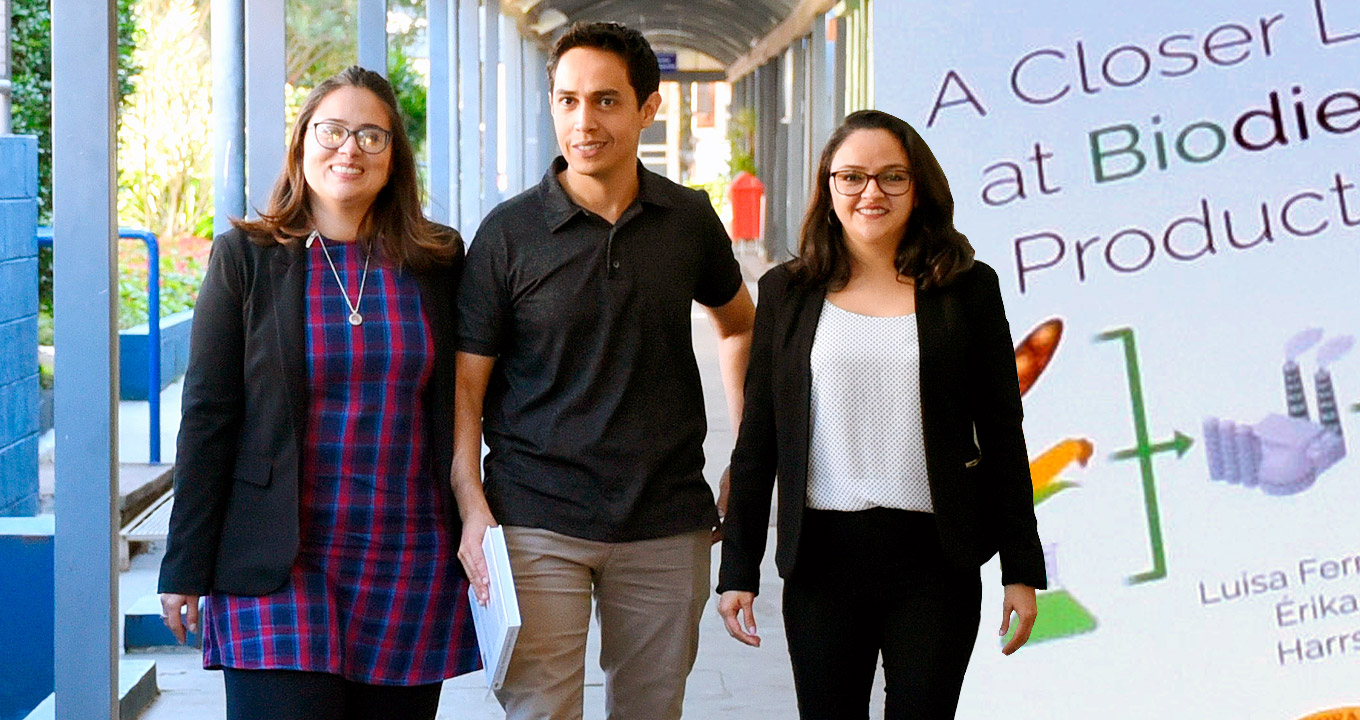 Da esq. para a dir., Érika Marques Takase, Harrson Santana e Luisa Fernanda Rios Pinto | Foto: Antoninho Perri Audiodescrição: em área coberta, imagem frontal e em plano médio, três pessoas em pé, uma ao lado da outra, sendo que duas delas olham para as lentes da câmara fotográfica. Eles estão em um extenso corredor, que segue às costas deles, sendo que à direita deles há uma ampla edificação, e à esquerda, colunas de concreto pintadas de azul, mureta e uma área externa com vegetação rasteira e árvores. À direita, ilustração em banner com frase em inglês closer look at biodiesel e três desenhos abaixo. Imagem 1 de 1