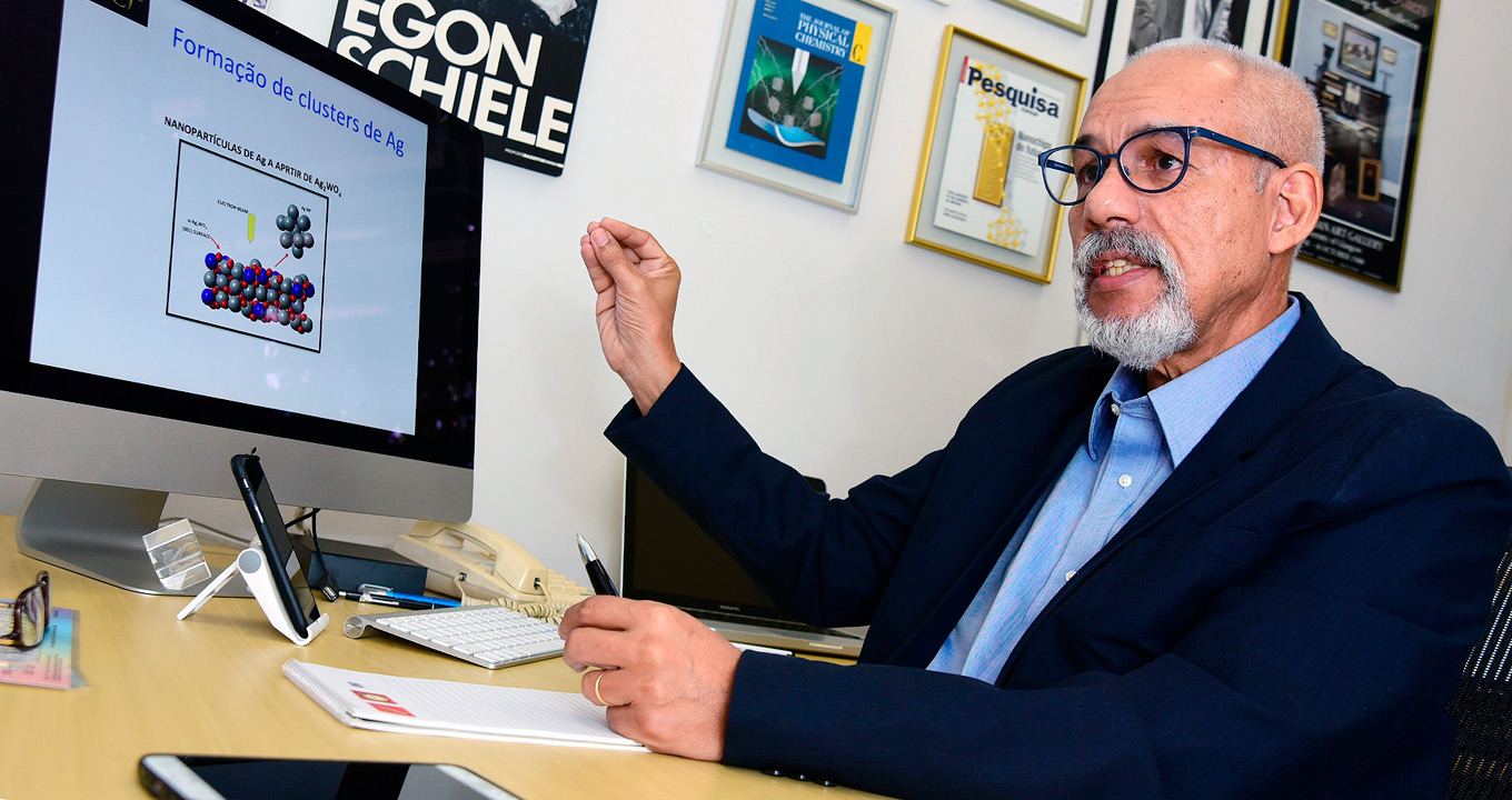 O professor Edison Zacarias da Silva, do IFGW-Unicamp | Foto: Antonio Scarpinetti Audiodescrição: em área interna, imagem em perspectiva e de busto, homem sentado em cadeira, com olhar voltado para a esquerda, fala e gesticula com a mão direita, mantendo o braço projetado para frente e as pontas do dedos unidas, apontando para um monitor de lcd que encontra-se à frente, sobre uma mesa de madeira. O monitor traz imagens de nanopartículas e o título formação de clusters de Ag. Ao lado dele, uma parede branca com quadros emoldurados com vidro de capas de revistas. Imagem 1 de 1.