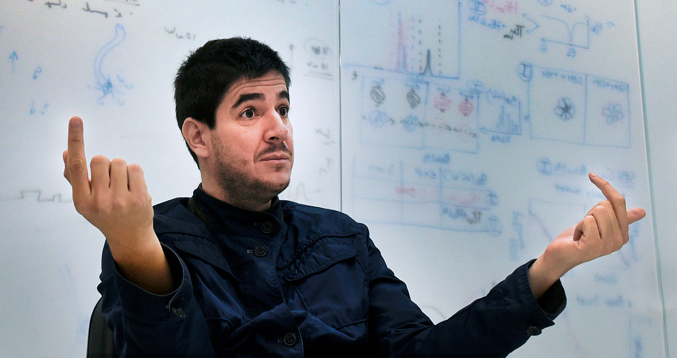 O professor Gustavo Wiederhecker, do Instituto de Física Gleb Wataghin | Foto: Antoninho Perri Audiodescrição: em sala de aula, imagem em perspectiva e de busto, homem sentado em cadeira, ao centro da imagem e com olhar voltado para a direita, sendo que ele gesticula com os braços, mantendo-os dobrados e projetados para frente, com as mão fechadas, e somente os dedos indicador e polegar voltados para cima. Às costas dele, em toda extensão da imagem, um tipo de lousa com fundo branco, anotações de fórmulas matemáticas e ilustrações. Ele usa camisa de mangas compridas na cor preta. Imagem 1 de 1.
